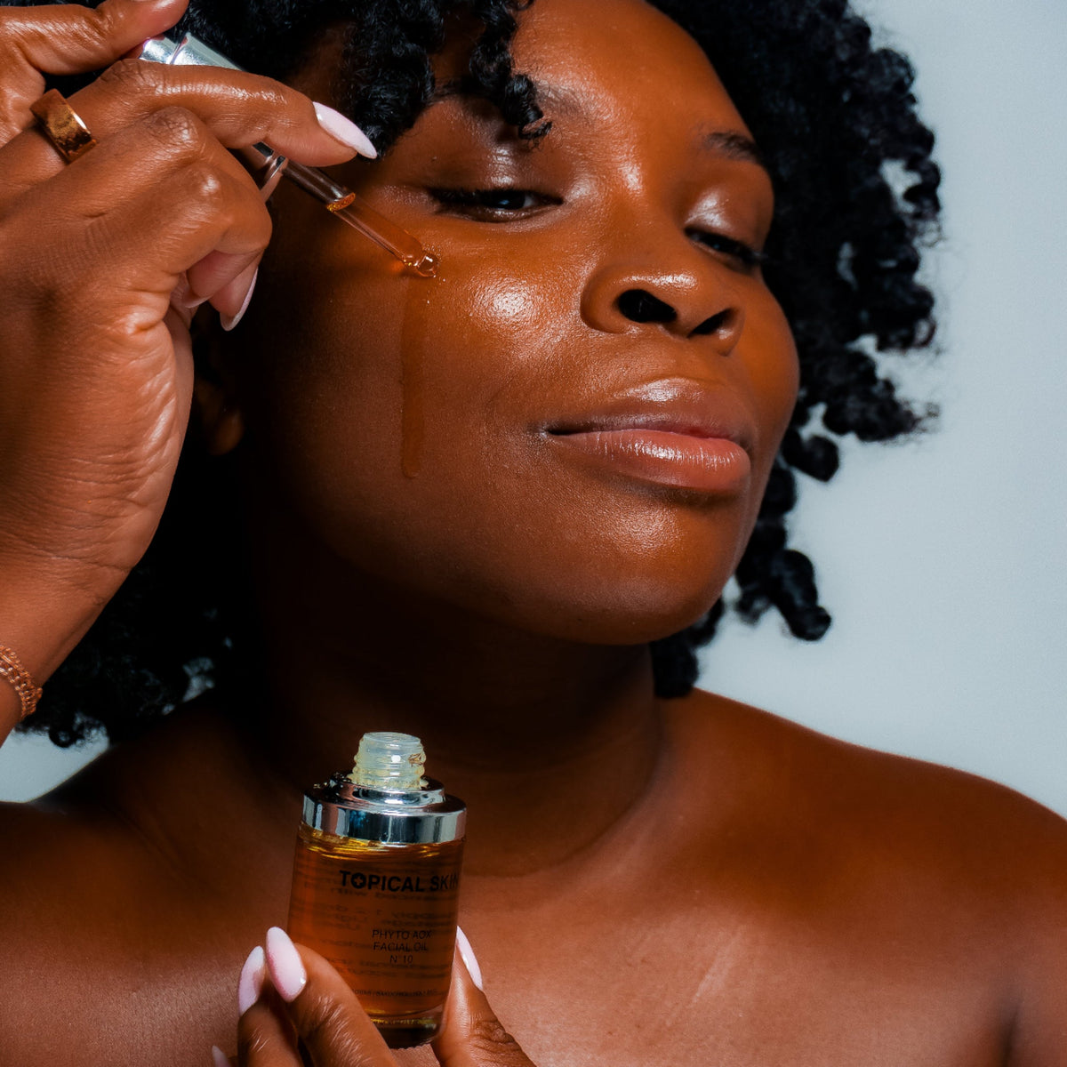 A woman with dark curly hair applies a golden-brown facial serum using a dropper, holding the bottle in her other hand against a light gray background.