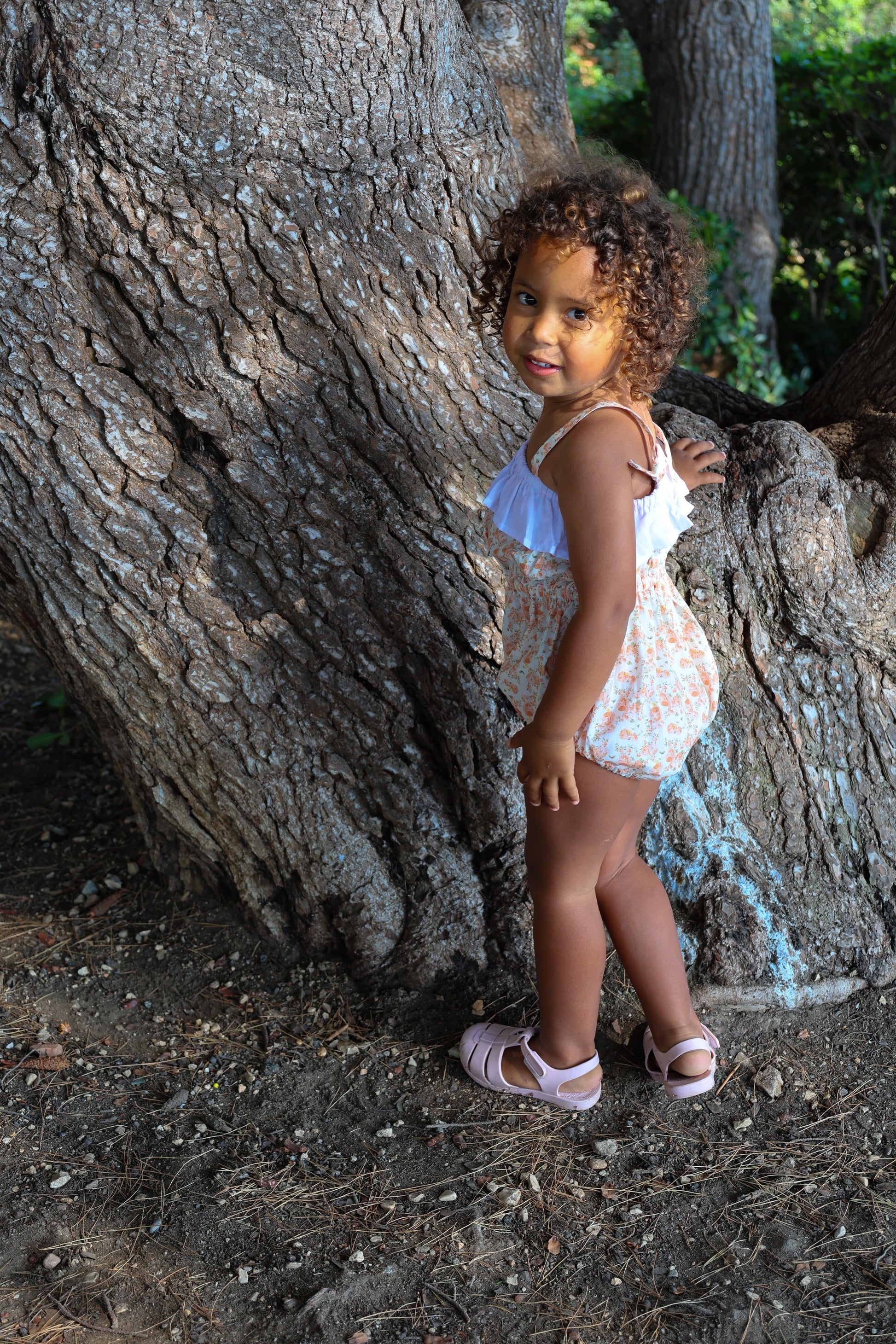 Young girl in a floral romper with adjustable straps standing next to a large tree