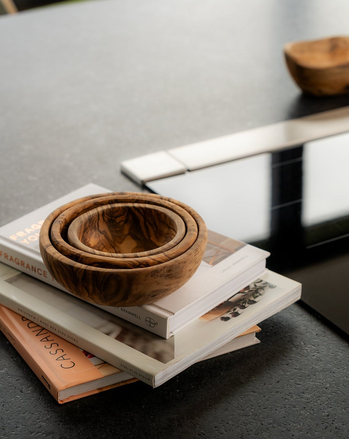 Three natural olive wood nesting bowls stacked on top of a pile of three books, creating a warm, rustic display on a tabletop.
