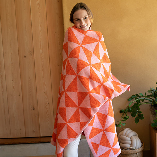 Woman wrapped in a neon orange and pink blanket with geometric patterns, standing in a room next to a plant and knotted pillow.