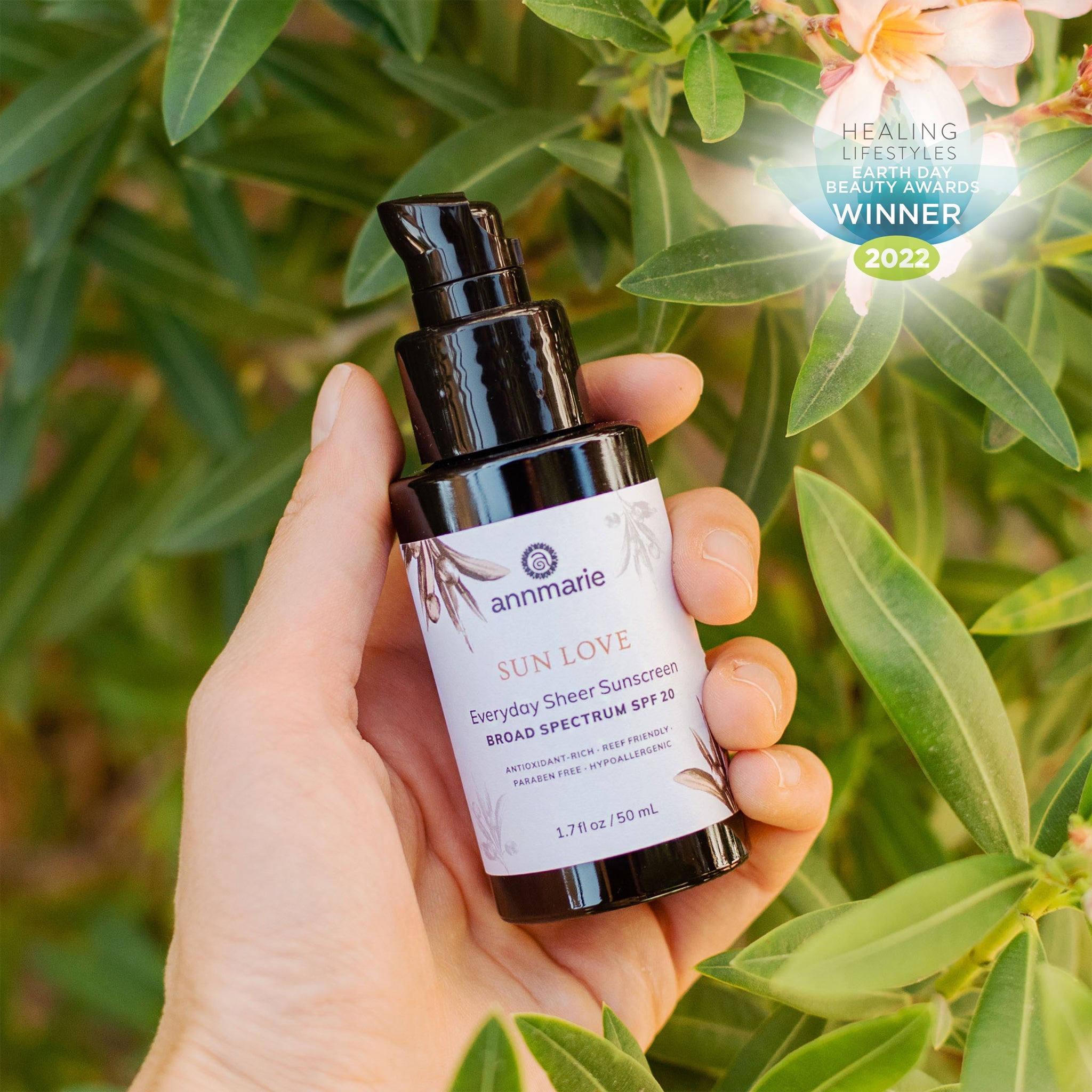 Hand holding Annmarie Sun Love Everyday Sheer Sunscreen SPF 20 bottle among green leaves with Earth Day Beauty Awards 2022 winner badge visible.