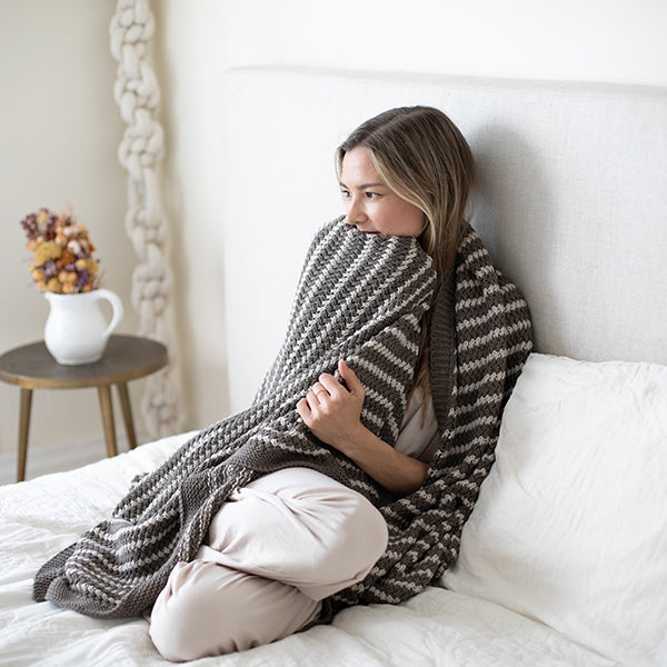 Woman sitting on a bed wrapped in a dark brown and white striped cotton blanket, with a side table and decorative items in the background.