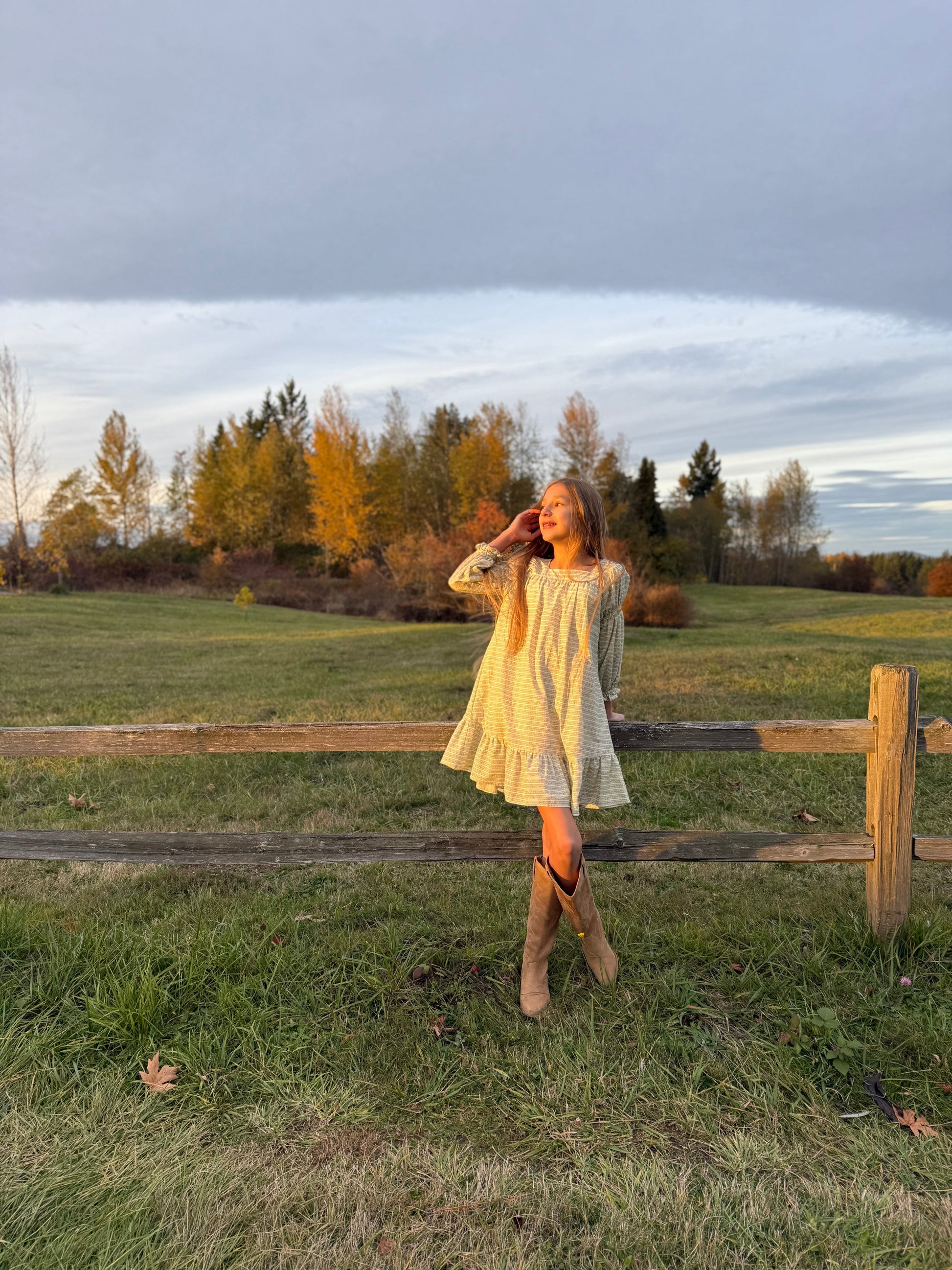 Woman in a yellow dress standing in a field with a wooden fence and trees in the background.