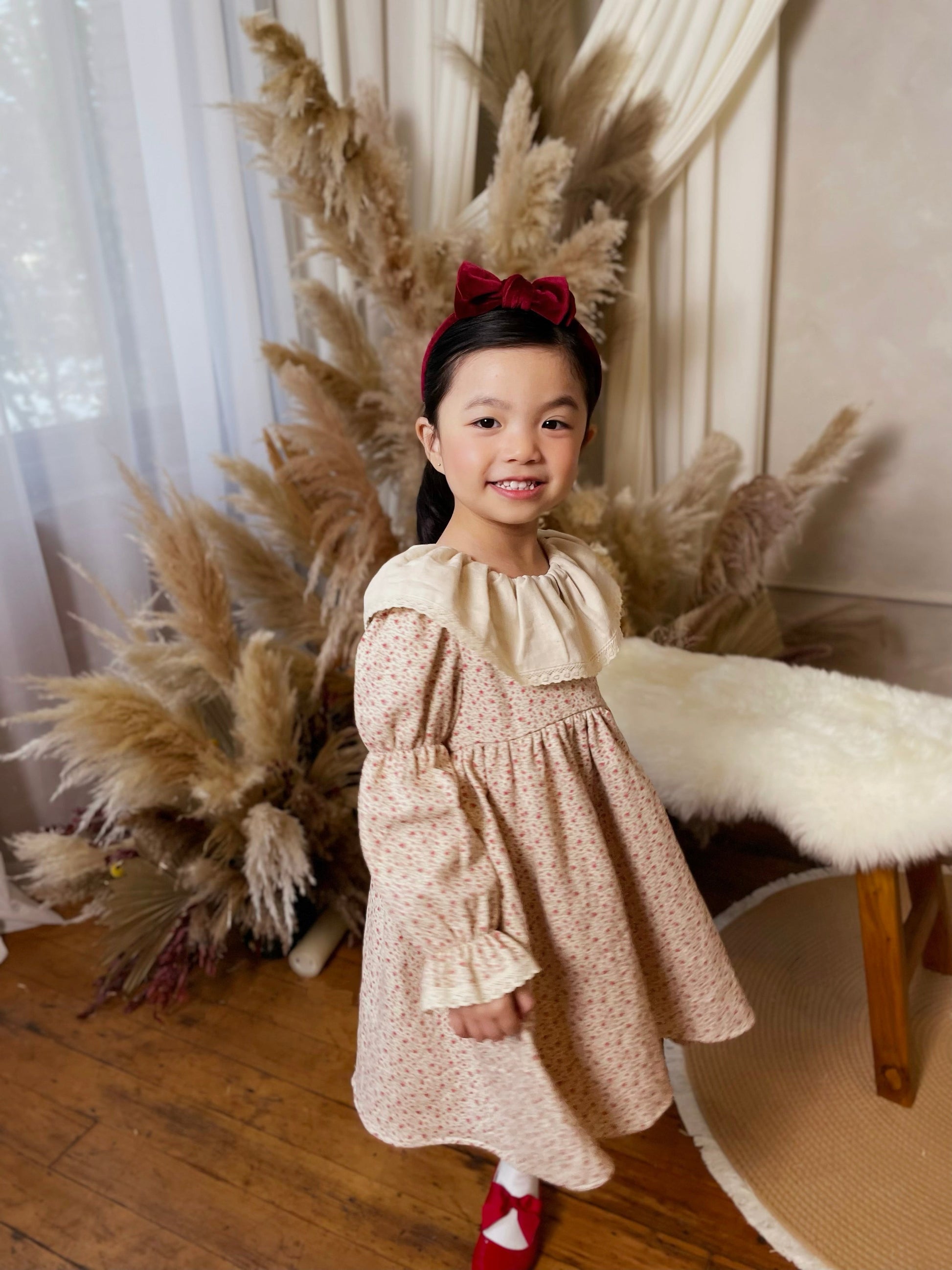 Young girl in a floral dress standing in front of decorative pampas grass and white curtains.