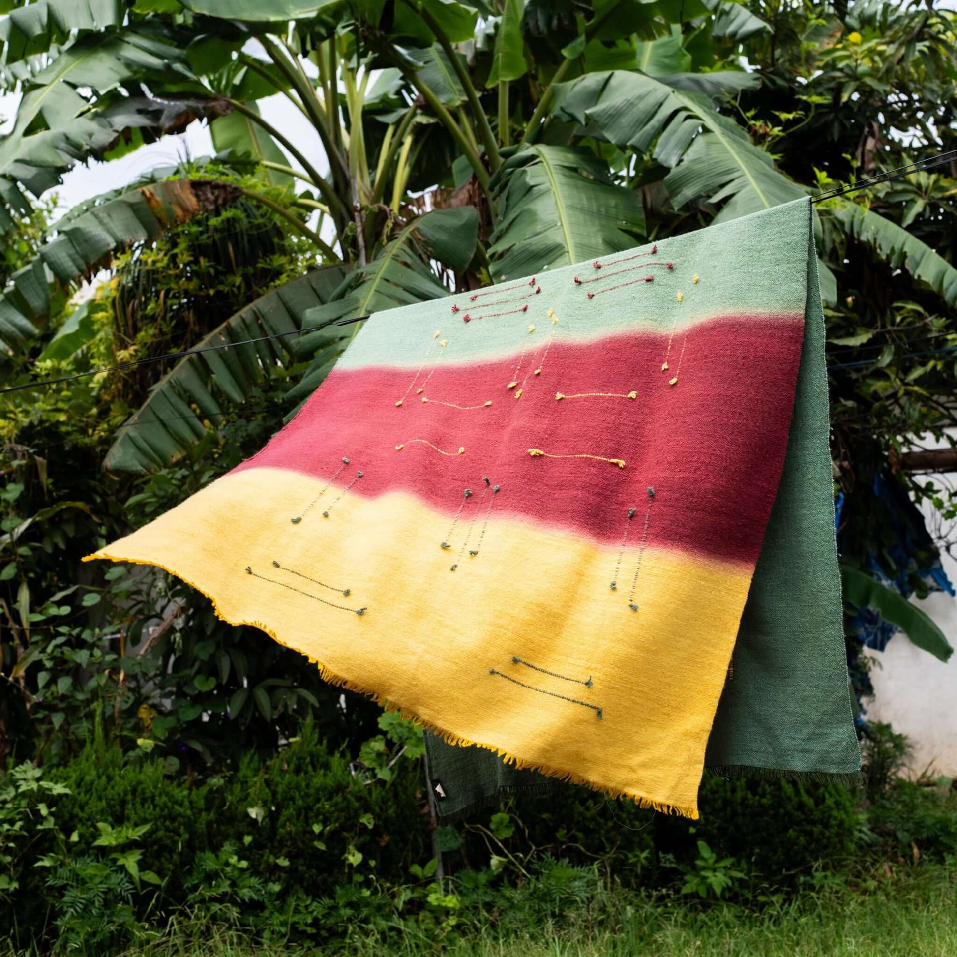Avocado Green Multi Color Warm Hues Hand Woven Hand Knotted Textured Merino Throw displayed outdoors among lush greenery.