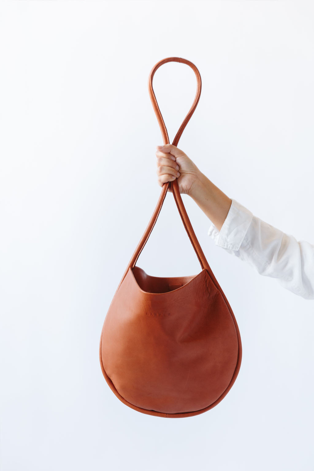 A woman's arm is visible, holding out a cognac leather shoulder bag by the thin strap. The leather is soft and shiny, and the bag is a half moon shape.