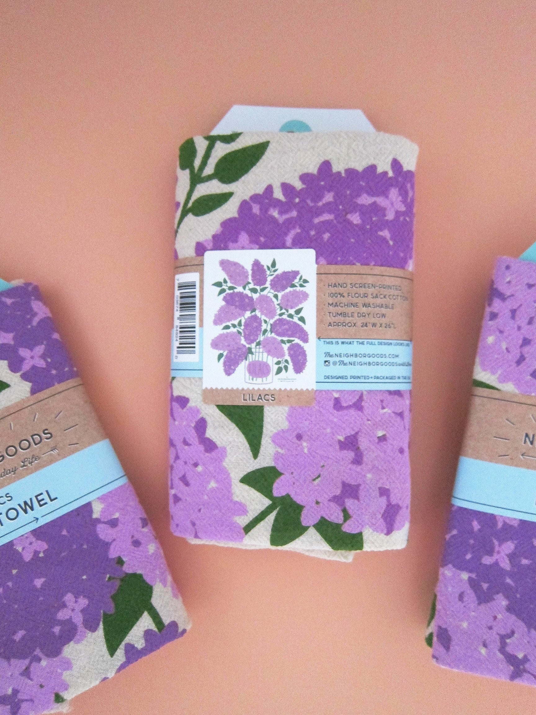 Back view of The Neighborgoods Lilacs dish towel in a ready-to-gift sleeve, showcasing product specifications and an image of the full tea towel.