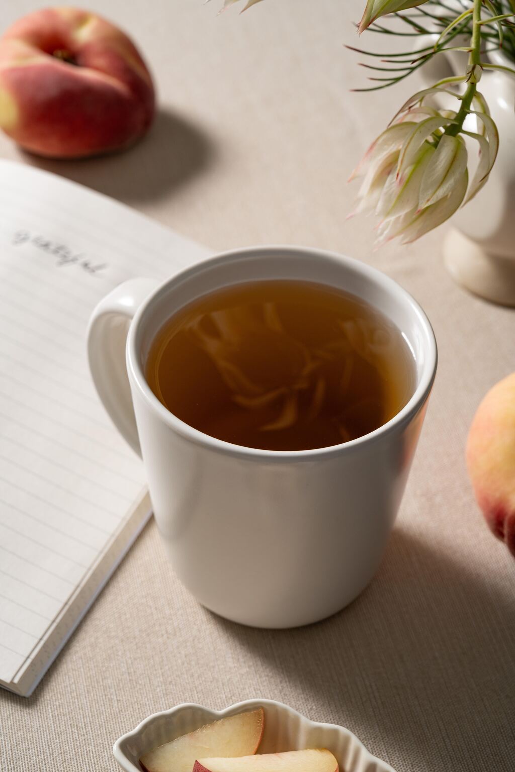 Off white ceramic Hamkke Home mug filled with tea beside peaches and an open journal on a neutral table.