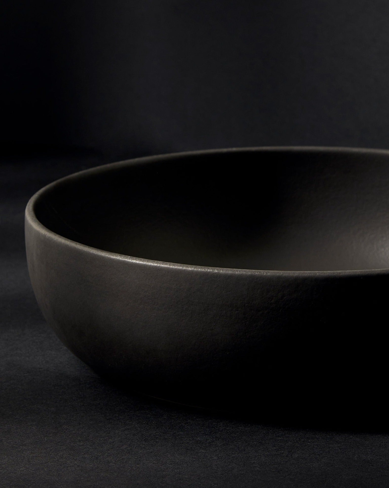 A matte black stoneware shallow serving bowl displayed against a dark background, showcasing its smooth, minimalist form and subtle texture.