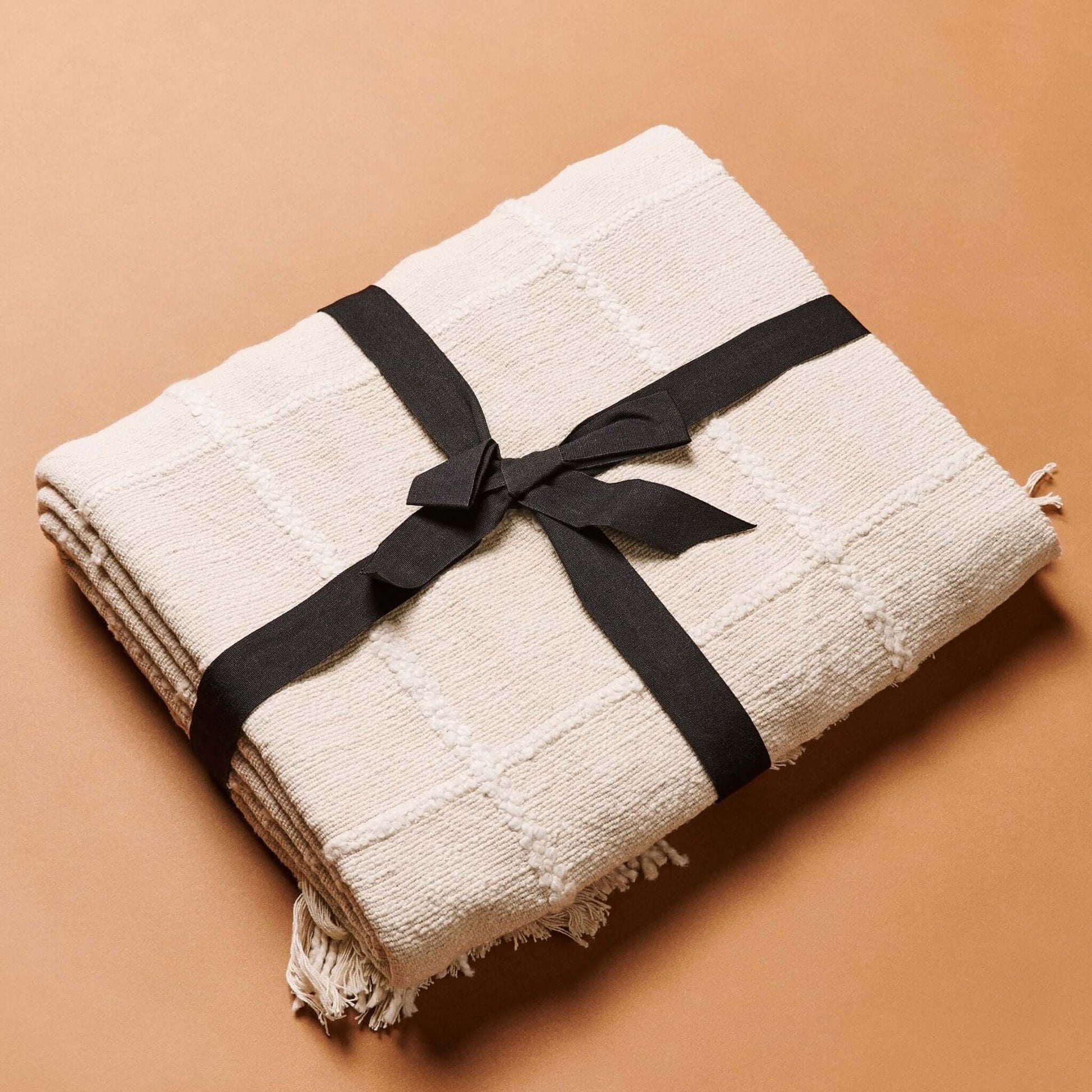 Sea Shell White Textured Checks Handloom Throw wrapped with a black ribbon on a tan background.
