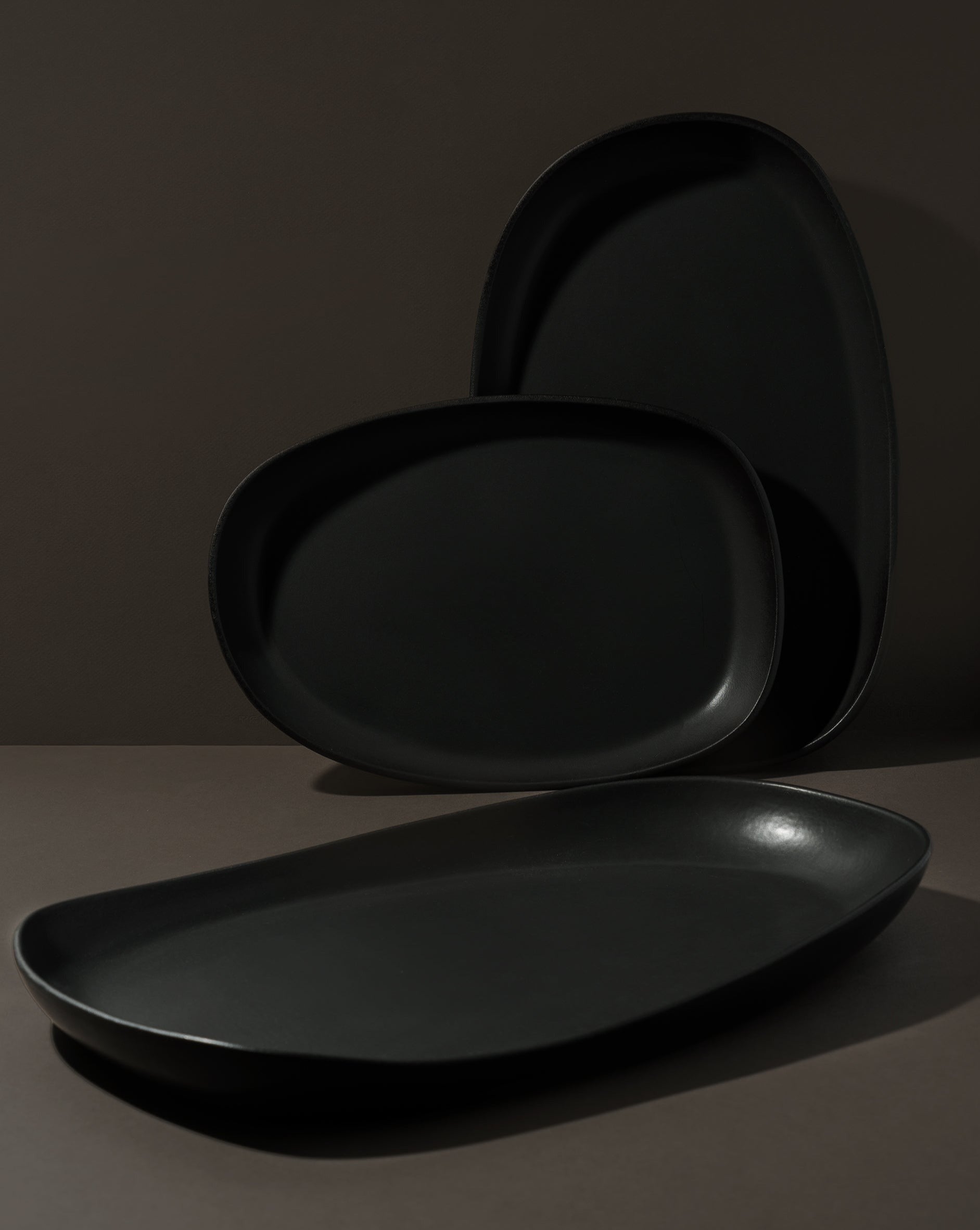 Matte black stoneware serving platter on a white background, with two additional platters standing beside it—one in landscape and one in portrait orientation—showcasing different angles of the elegant, handcrafted design.