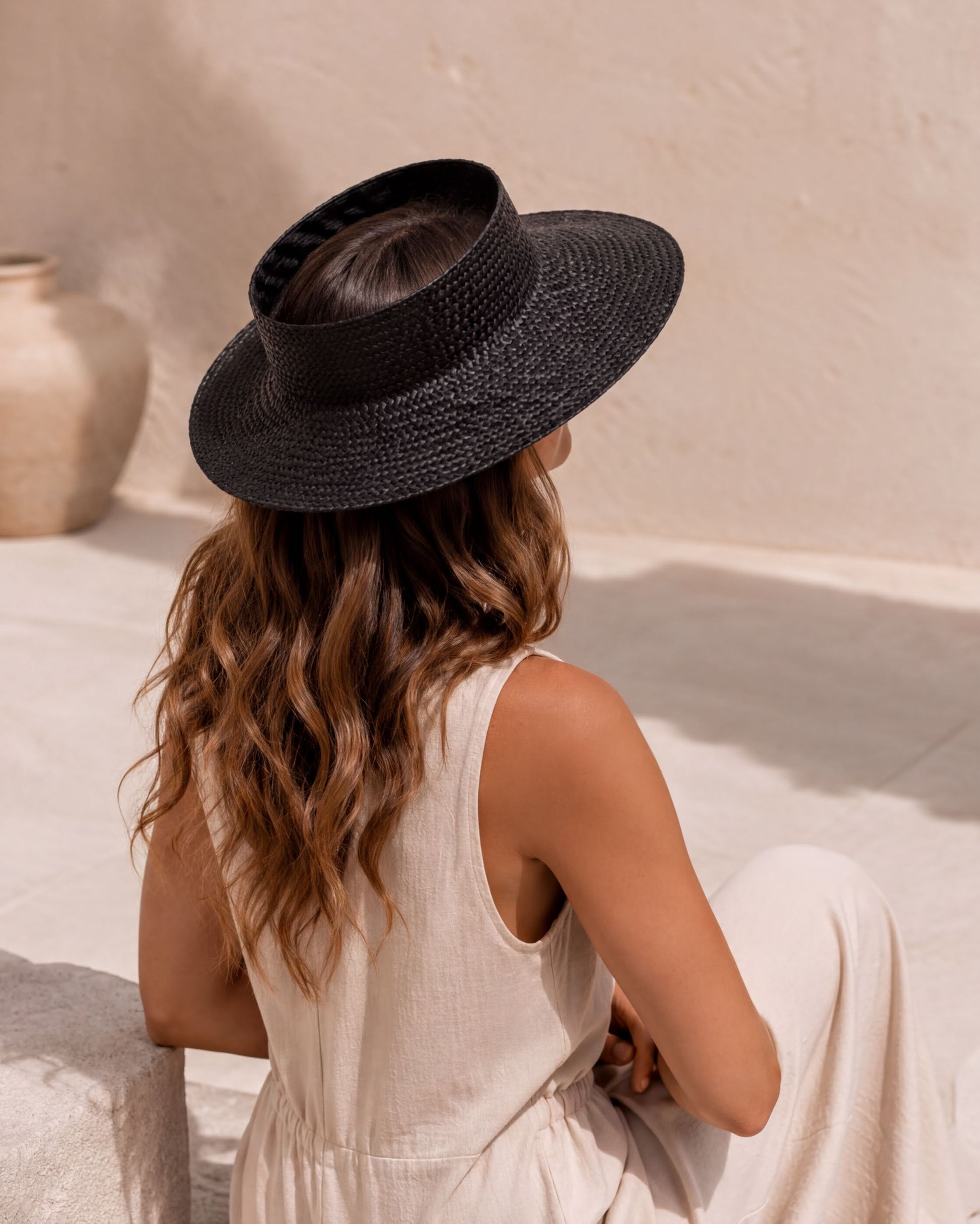 A woman wearing a black hat sits on a white surface, facing away from the camera.