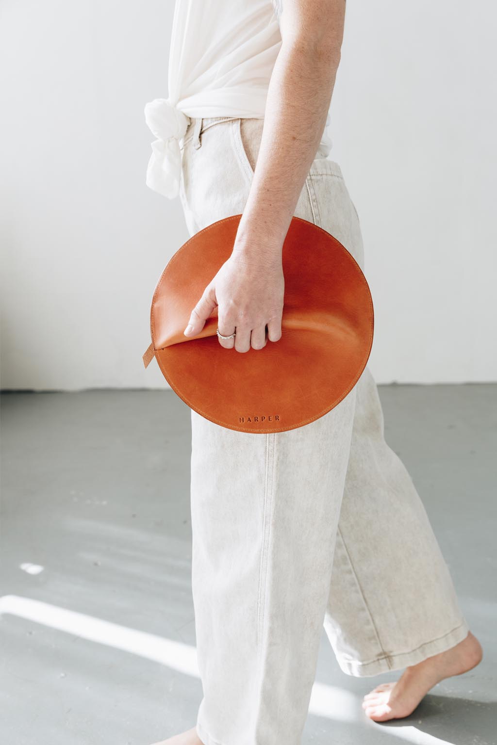 A woman holds a circular cognac brown leather clutch in her hands as she walks.