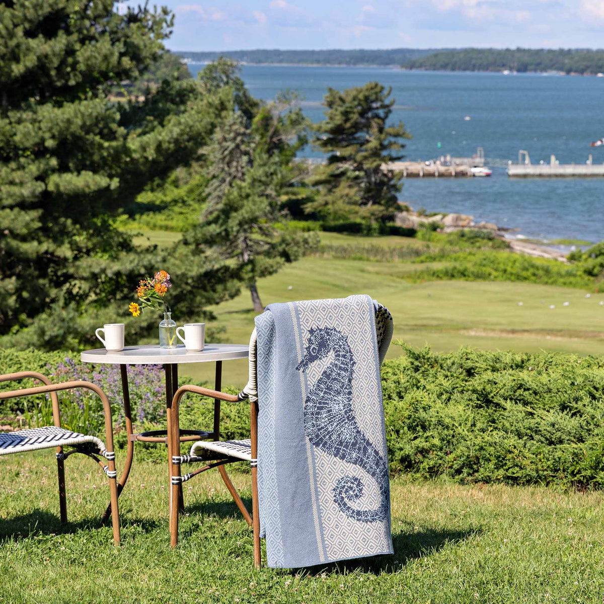An outdoor table with two chairs is set on grass overlooking water, trees, and a distant pier. Two mugs and a small vase of flowers decorate the table, while an In2Green Seahorse Square - Cotton Sustainable Knit Throw Blanket | Coastal is draped over one seat.