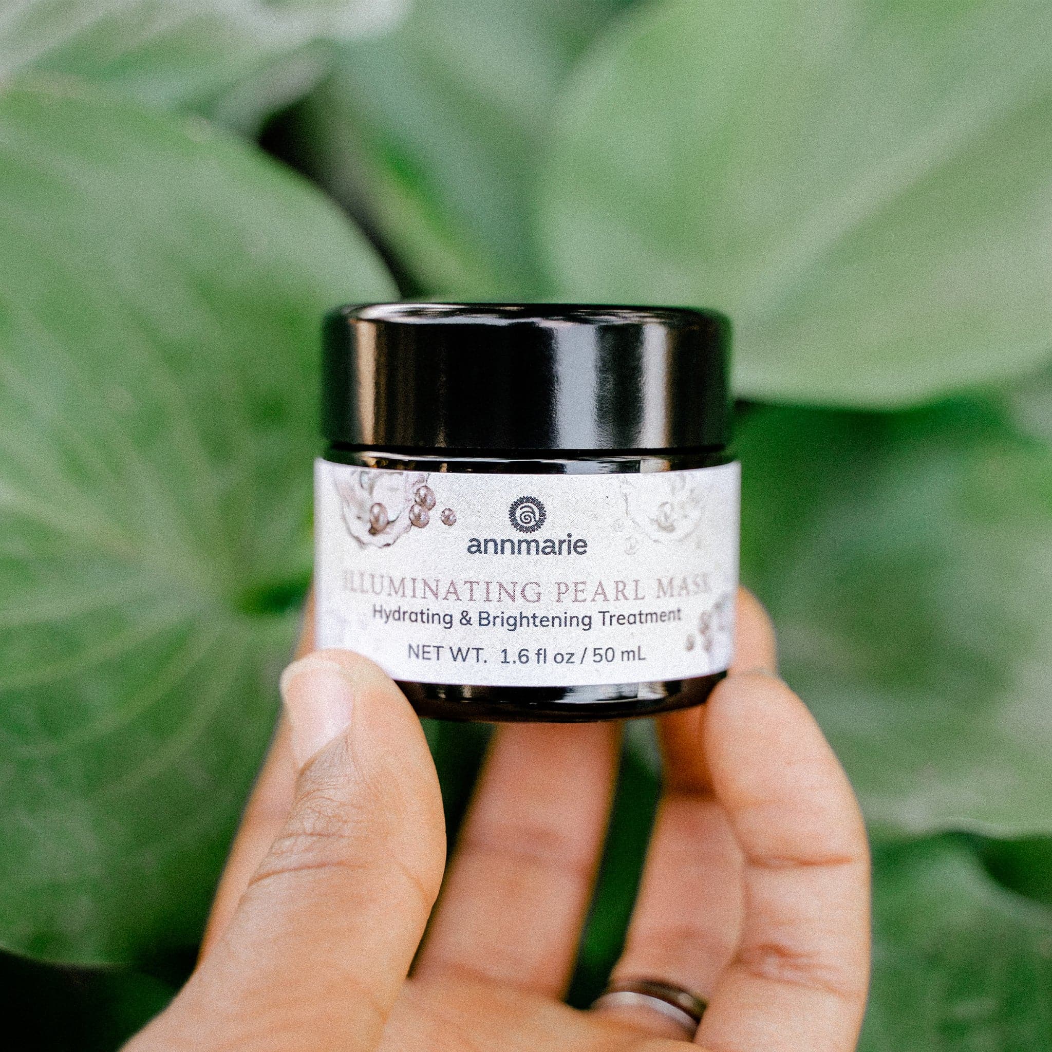 Hand holding a jar of Annmarie Skin Care Illuminating Pearl Mask against a green leafy background