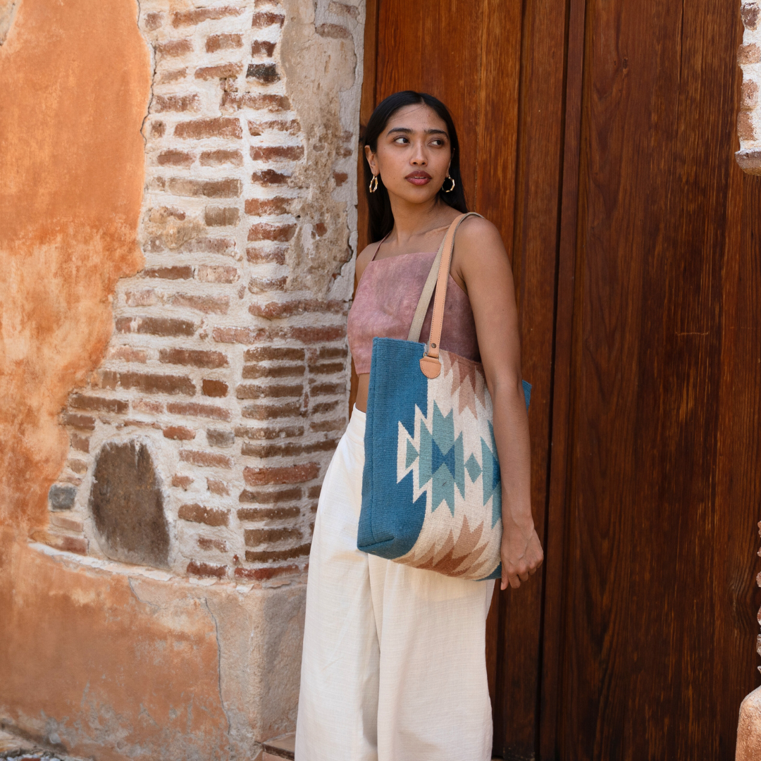 Woman carrying handwoven bucket bag in rose and blue with abstract diamond and lightning pattern, leather shoulder straps, and relaxed shape, ideal for everyday or beach use.
