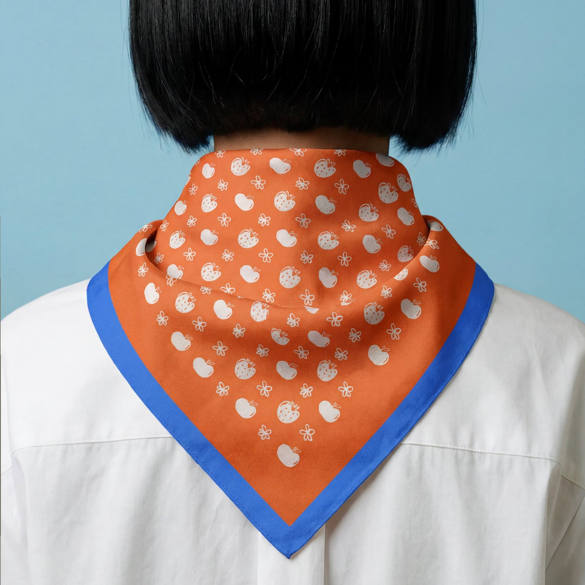woman wearing an orange scarf with white apples, strawberries and flowers pattern, with blue border on a blue background