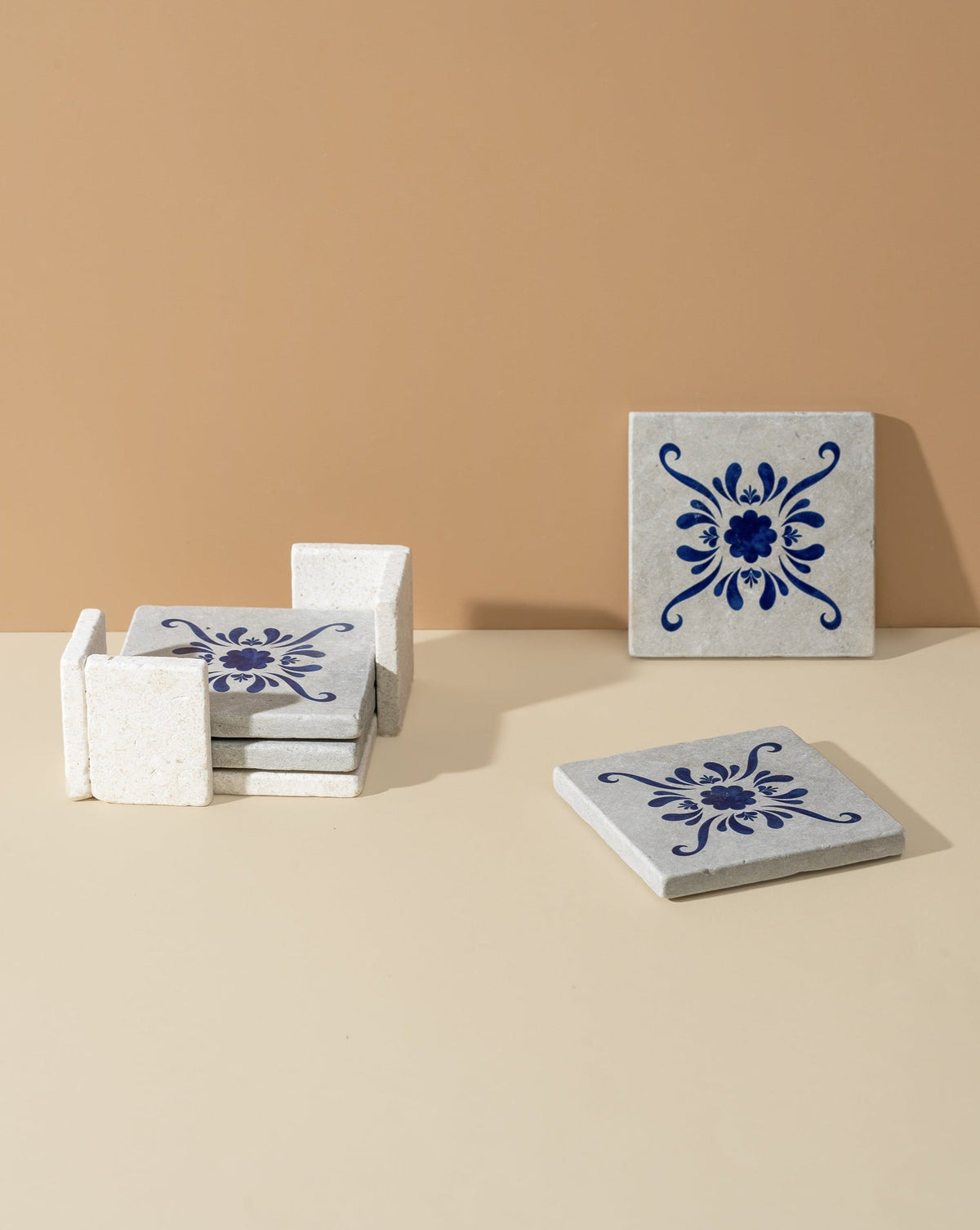 A set of four square white marble coasters with blue floral patterns, displayed on a beige background. Three are stacked on the side, while two are laid out separately.