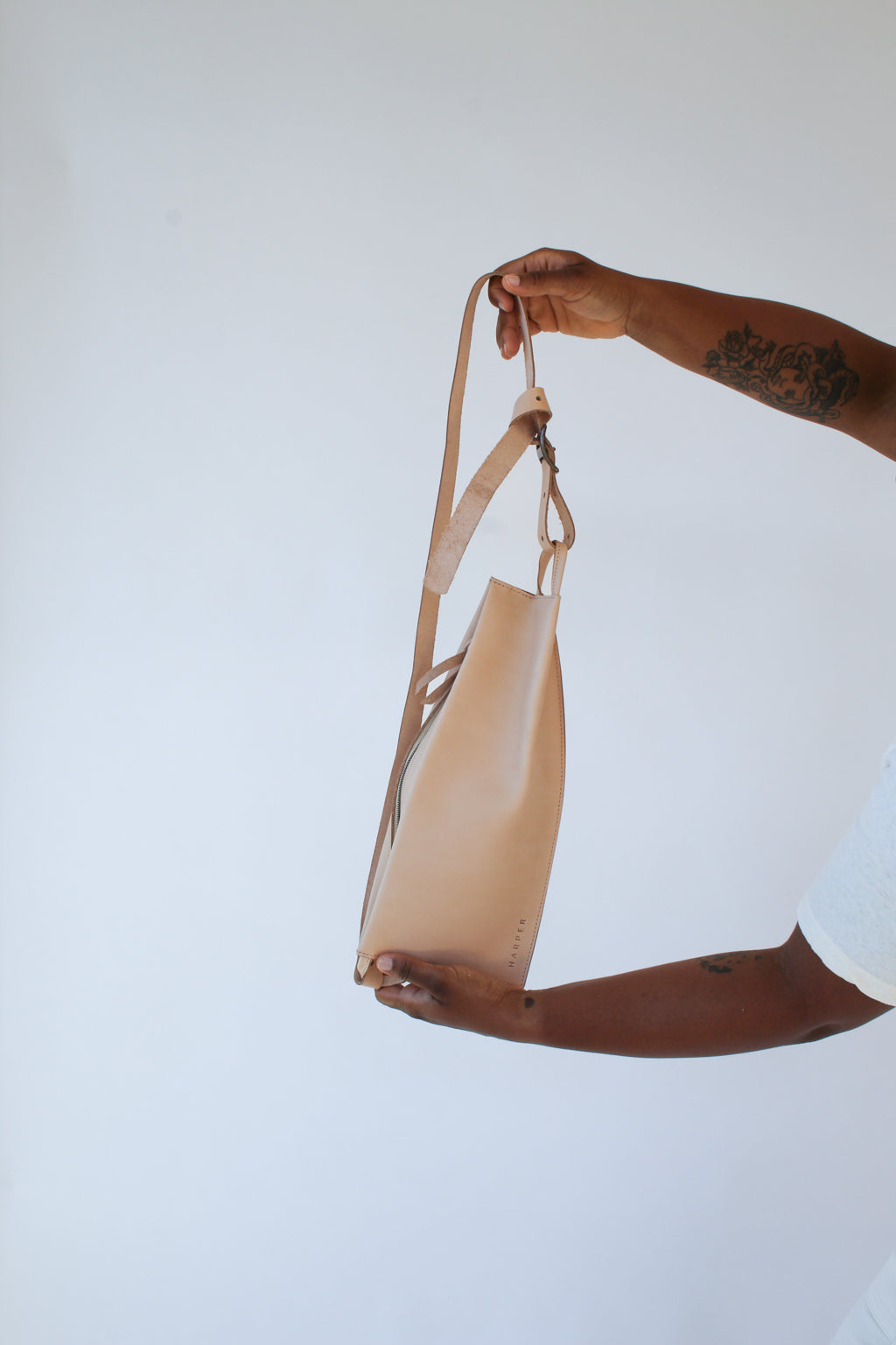 A woman holds out a natural, undyed leather fanny pack shoulder bag against a white background.