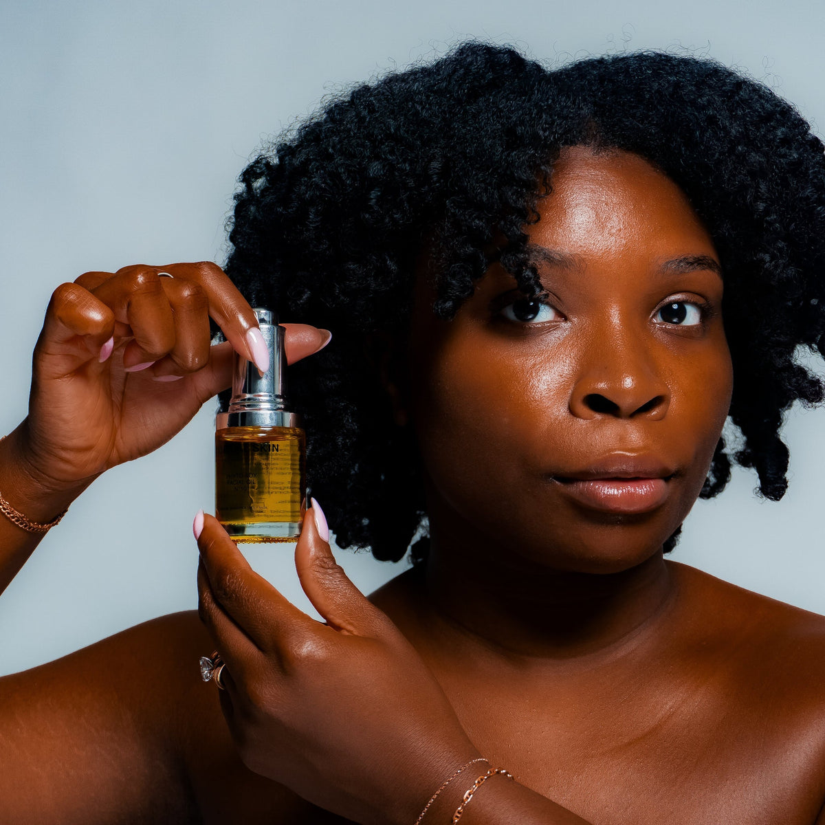 A woman with curly black hair holds a small glass bottle of golden-yellow oil against a light gray background.
