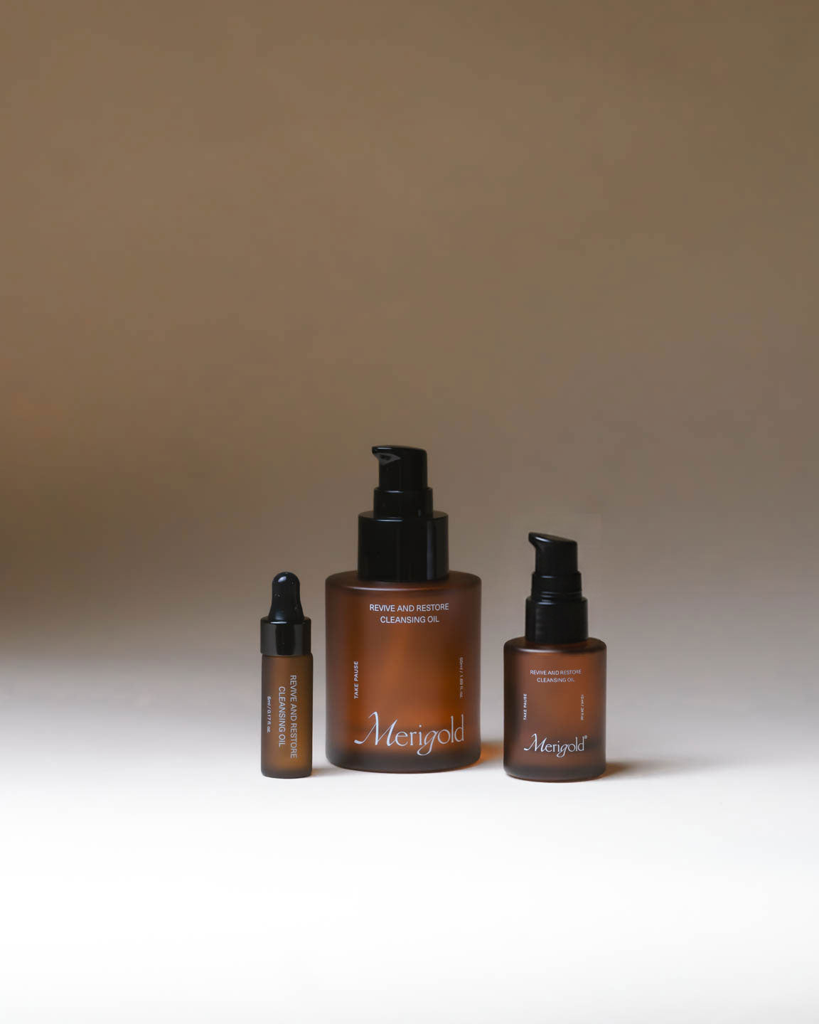 Merigold Revive and Restore Cleansing Oil in three sizes, displayed in amber glass bottles with black pumps on a neutral background.