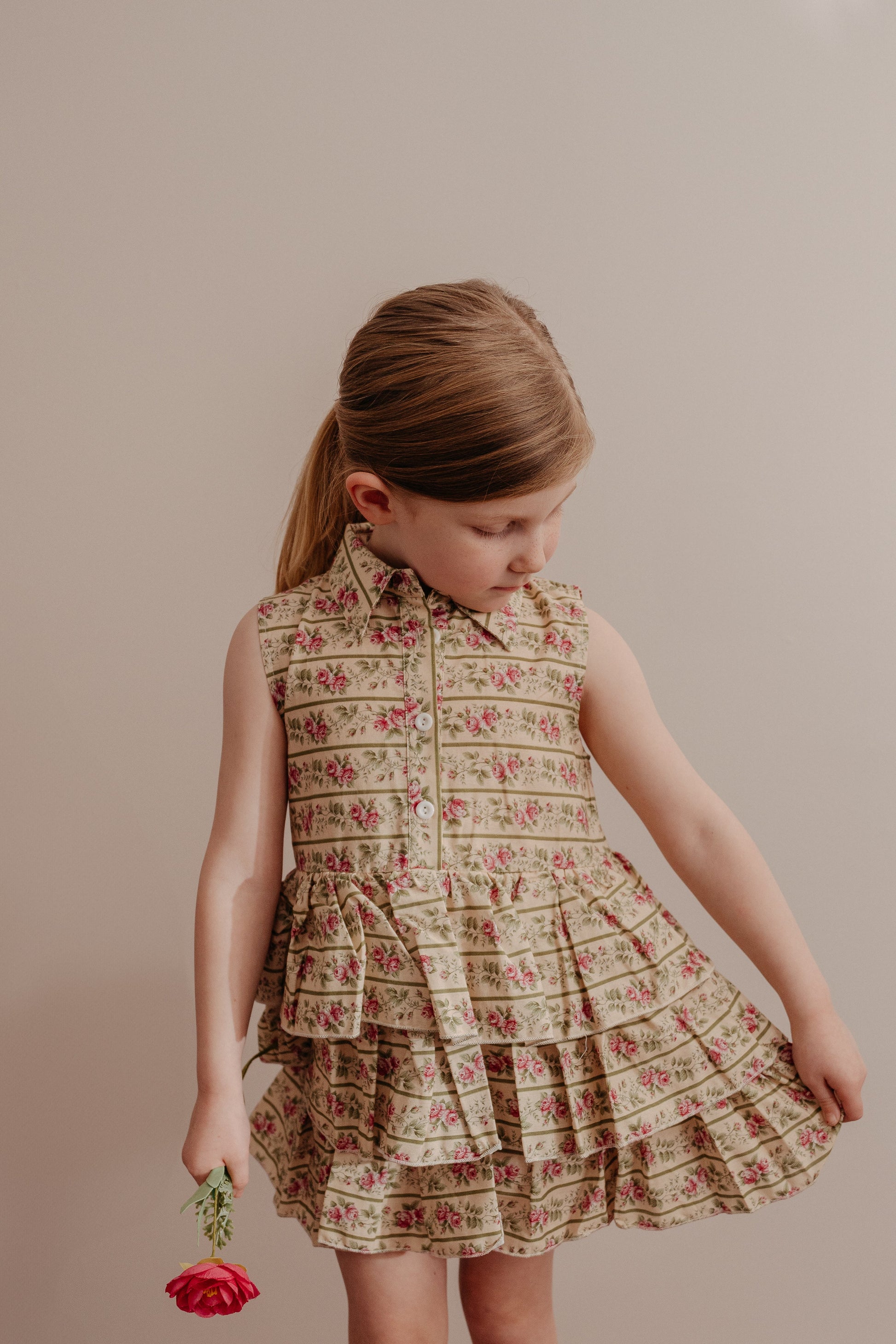 A young child wearing a sleeveless, floral print dress with a button-down front and a collar, featuring a multi-tiered ruffle skirt.