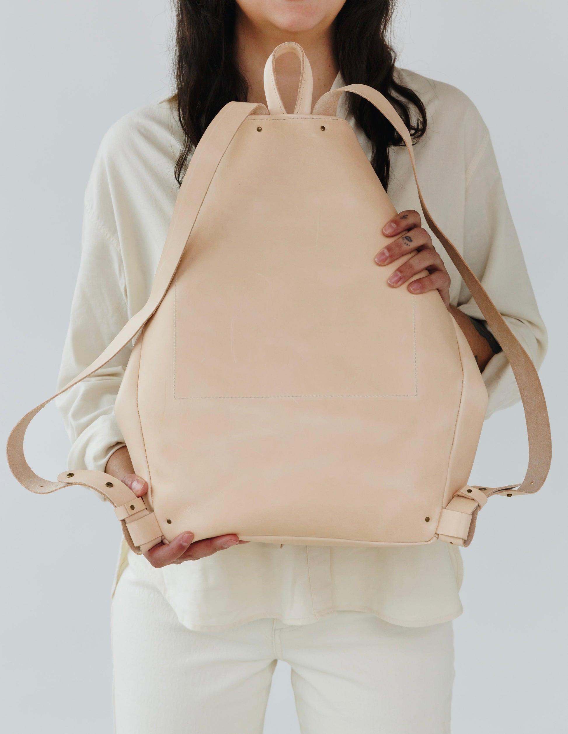A woman holds out a natural, undyed leather backpack to show its reverse side.