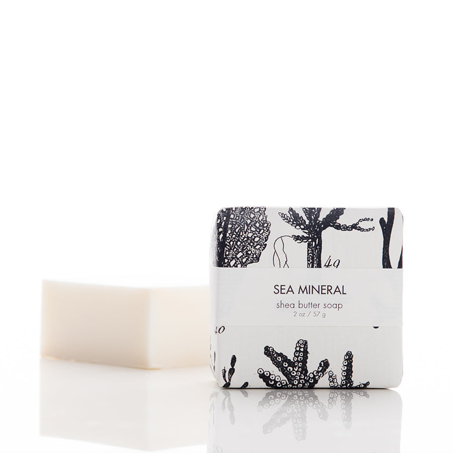 A wrapped bar of sea mineral shea butter soap is positioned next to an unwrapped, identical bar. The packaging features black and white illustrations of sea plants. The label reads "SEA MINERAL shea butter soap 2 oz / 57 g." The background is white and reflective.