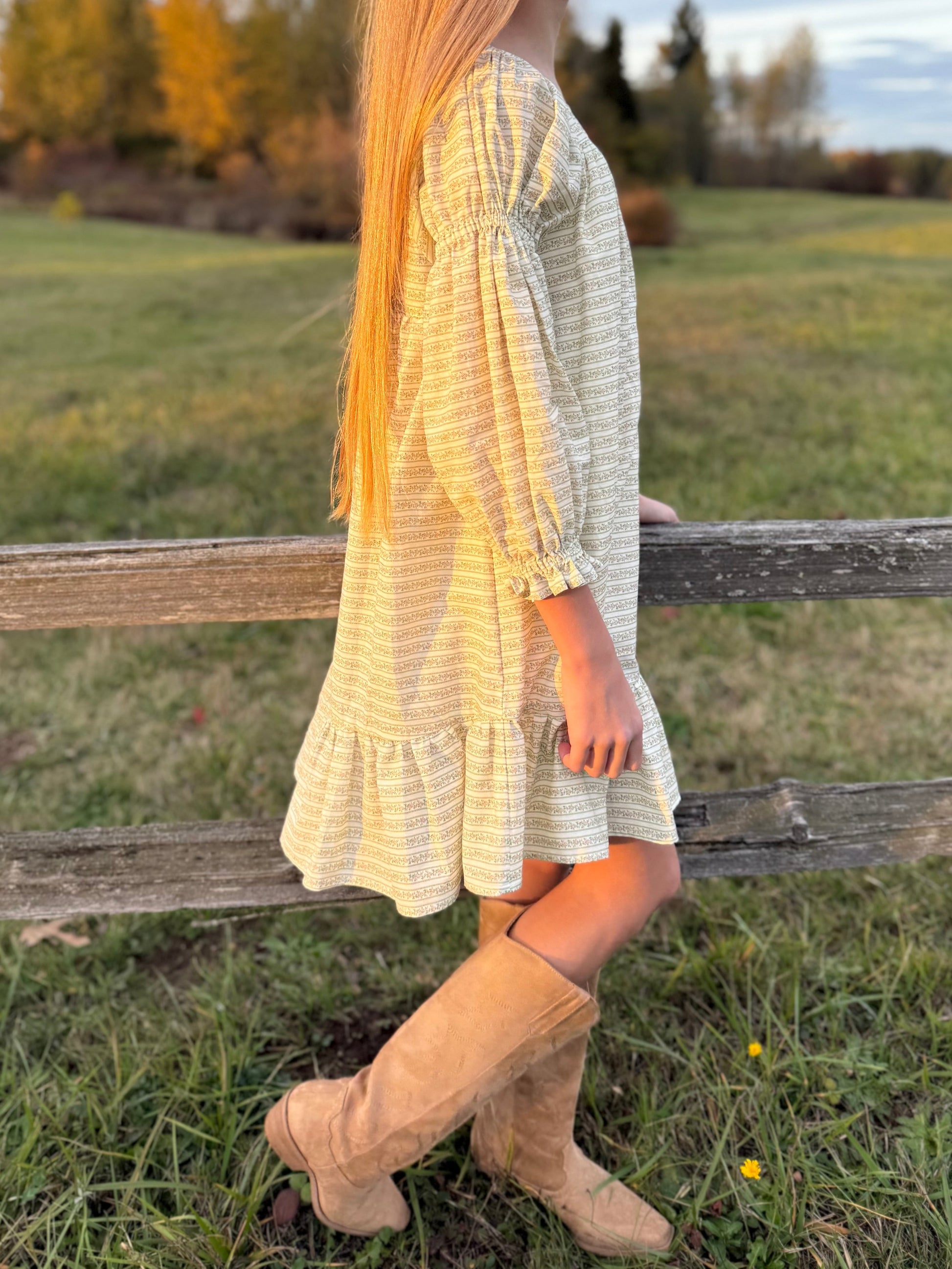 Person wearing a light dress and beige boots standing near a wooden fence in an outdoor setting.