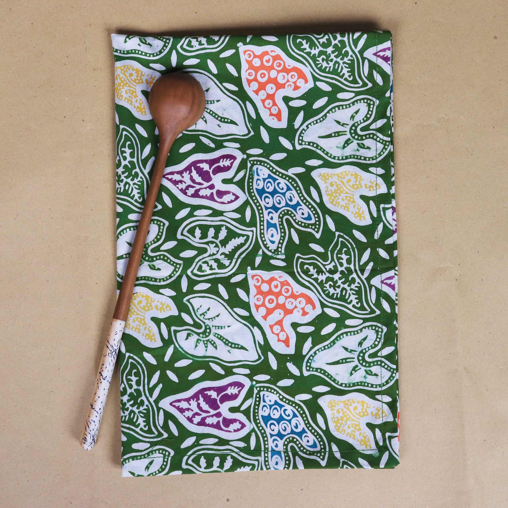 Kitchen tea towel with colorful leaves pattern on a green background, accompanied by a wooden spoon.