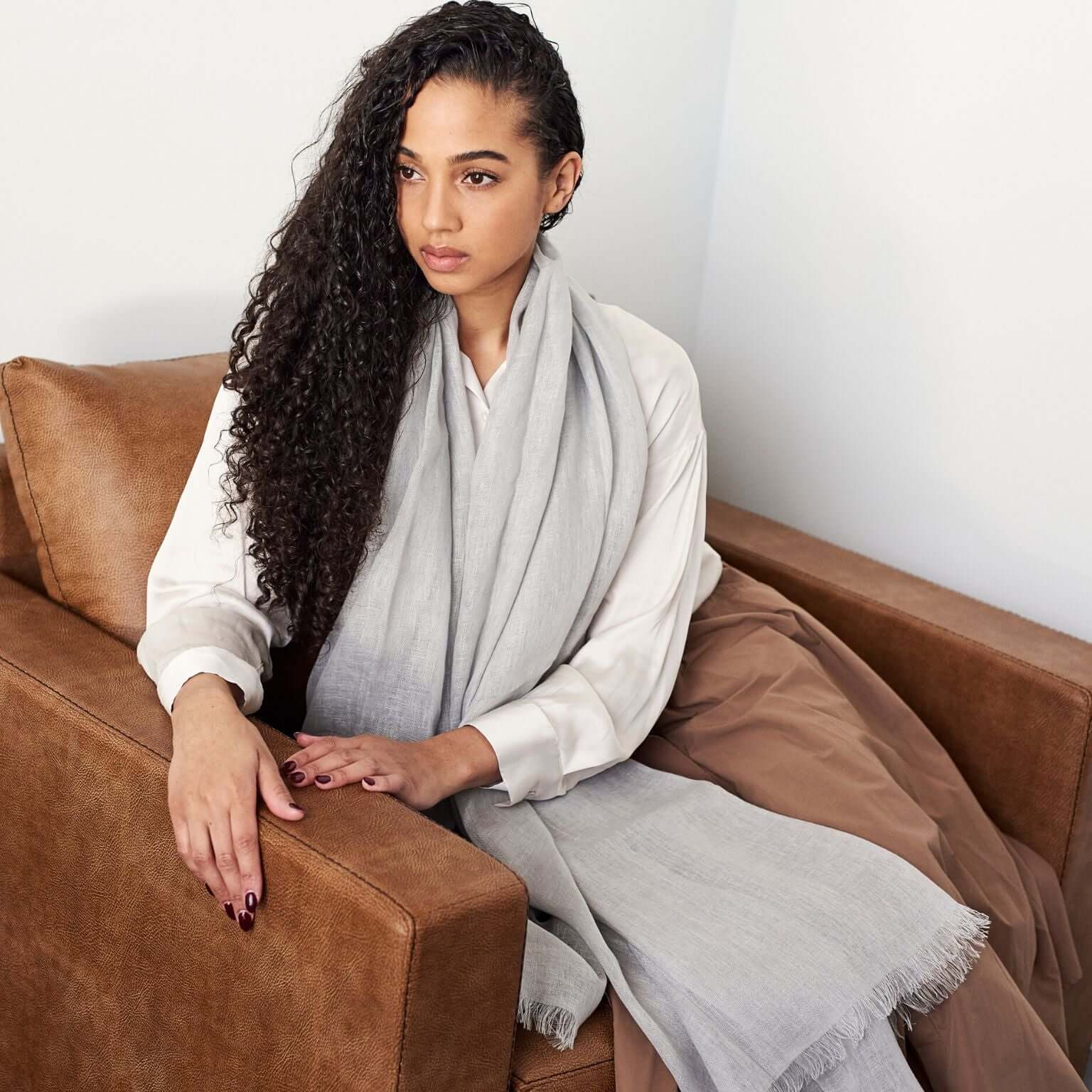 Woman wearing a Pebble Light Gray Hand Woven Soft Linen Scarf, sitting comfortably on a brown couch.
