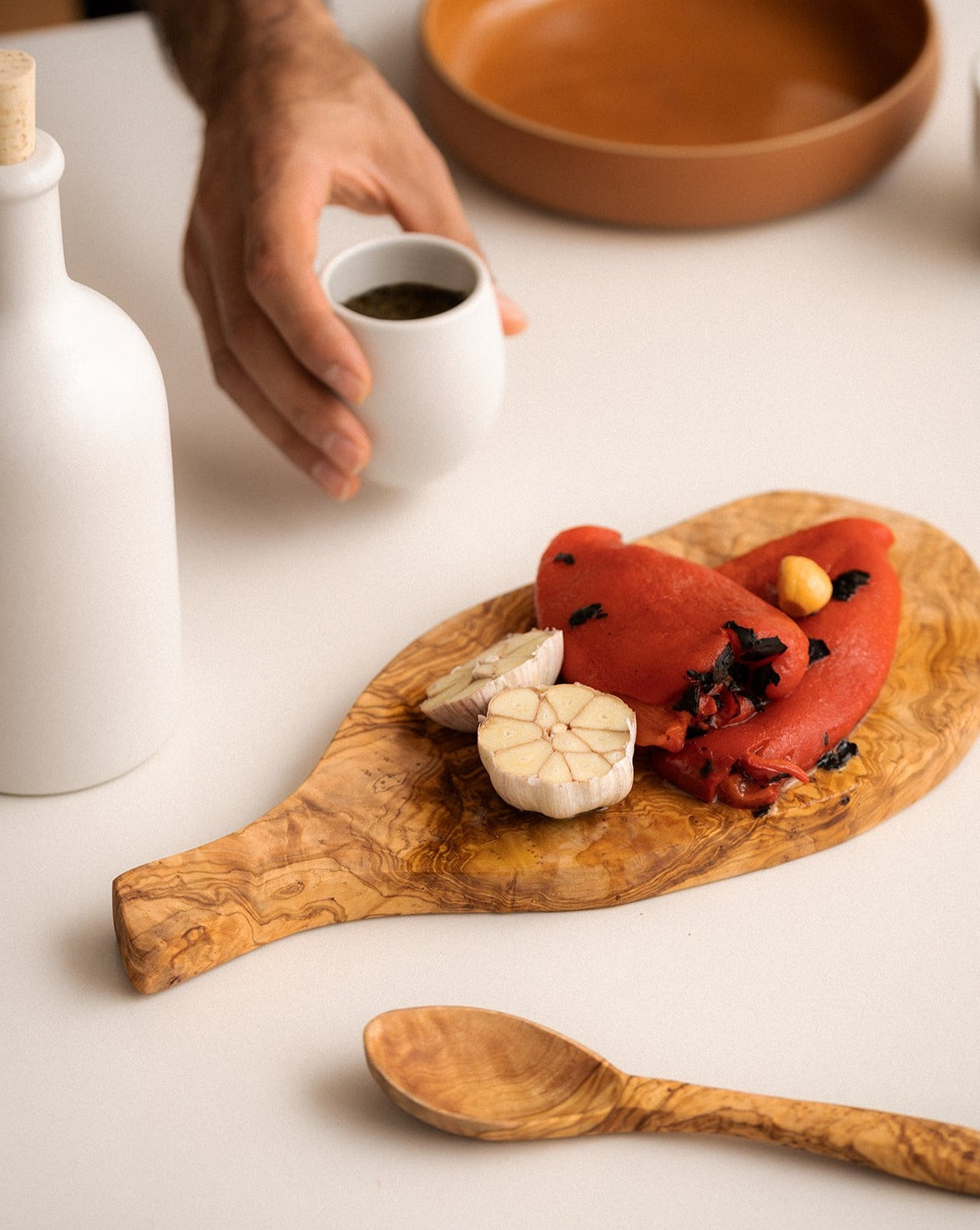 A natural olive wood cheese board with an organic shape, now topped with assorted food items. Beside it are a small wooden spoon, a tiny cup, and an olive oil dispenser, all arranged on a neutral surface for a cozy, rustic look.

