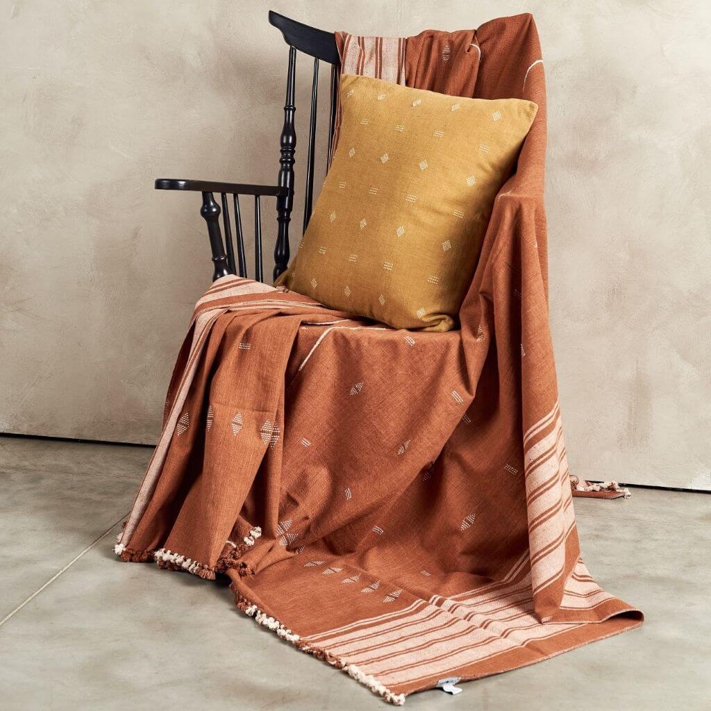 Nira Ochre Hand Spun Organic Cotton Handloom Pillow on a chair with woven throw blanket.