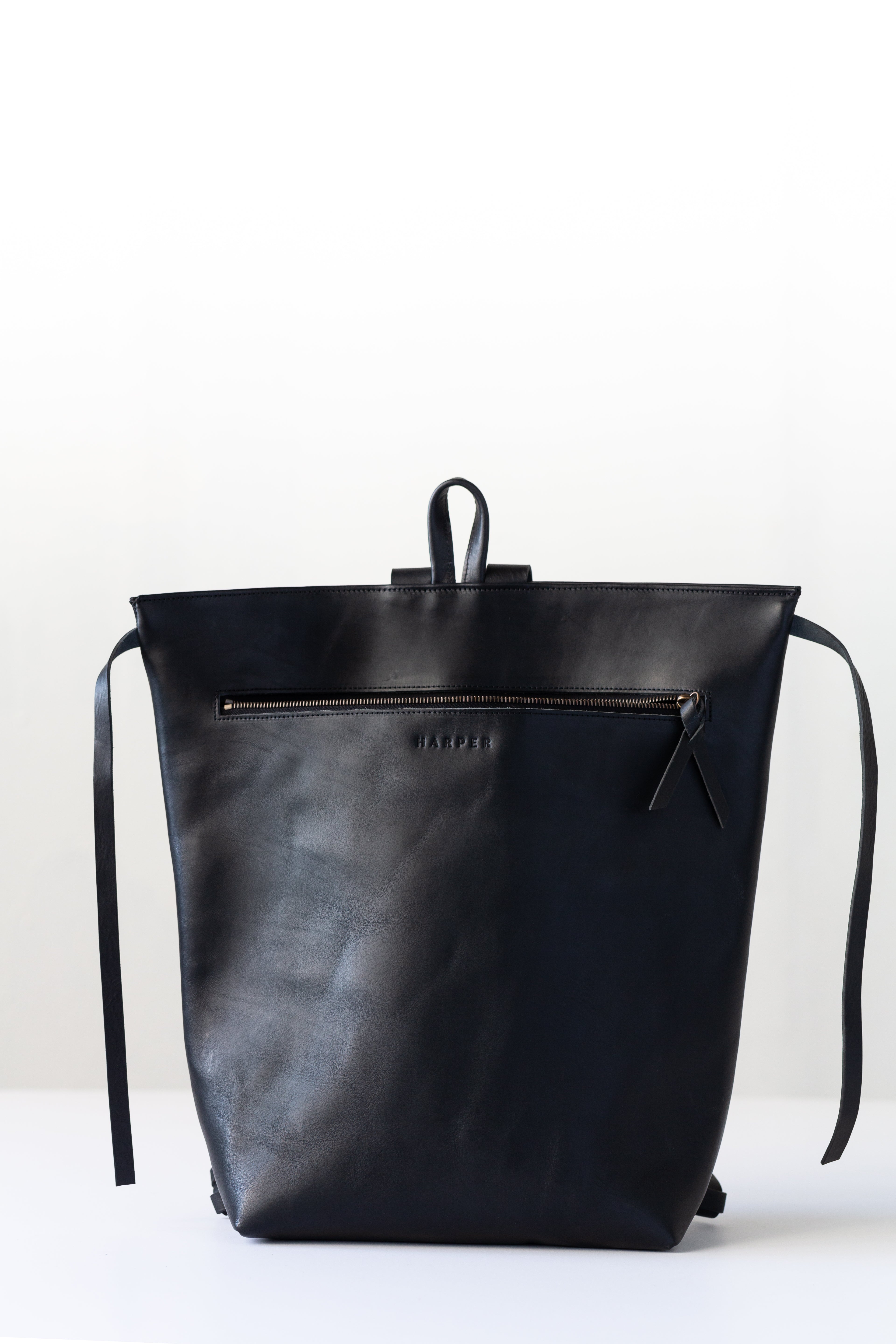 A woman holds out her arm, which is looped through the shoulder straps of a black leather backpack. The top corners of the backpack are cinched with straps that knot over a zipper, creating a unique shape.