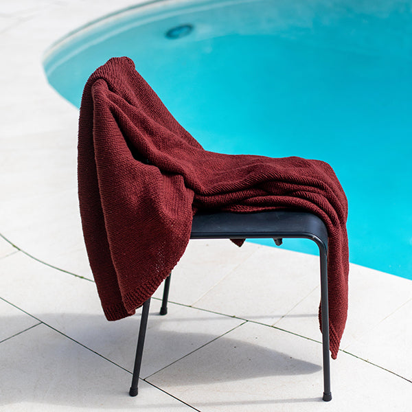 Red organic cotton throw draped over an outdoor chair by a swimming pool.