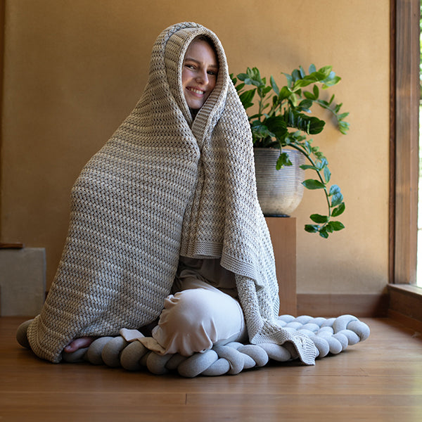 Person wrapped on top of a knotted floor cushion, wearing a silver cotton throw blanket with a plant in the background.