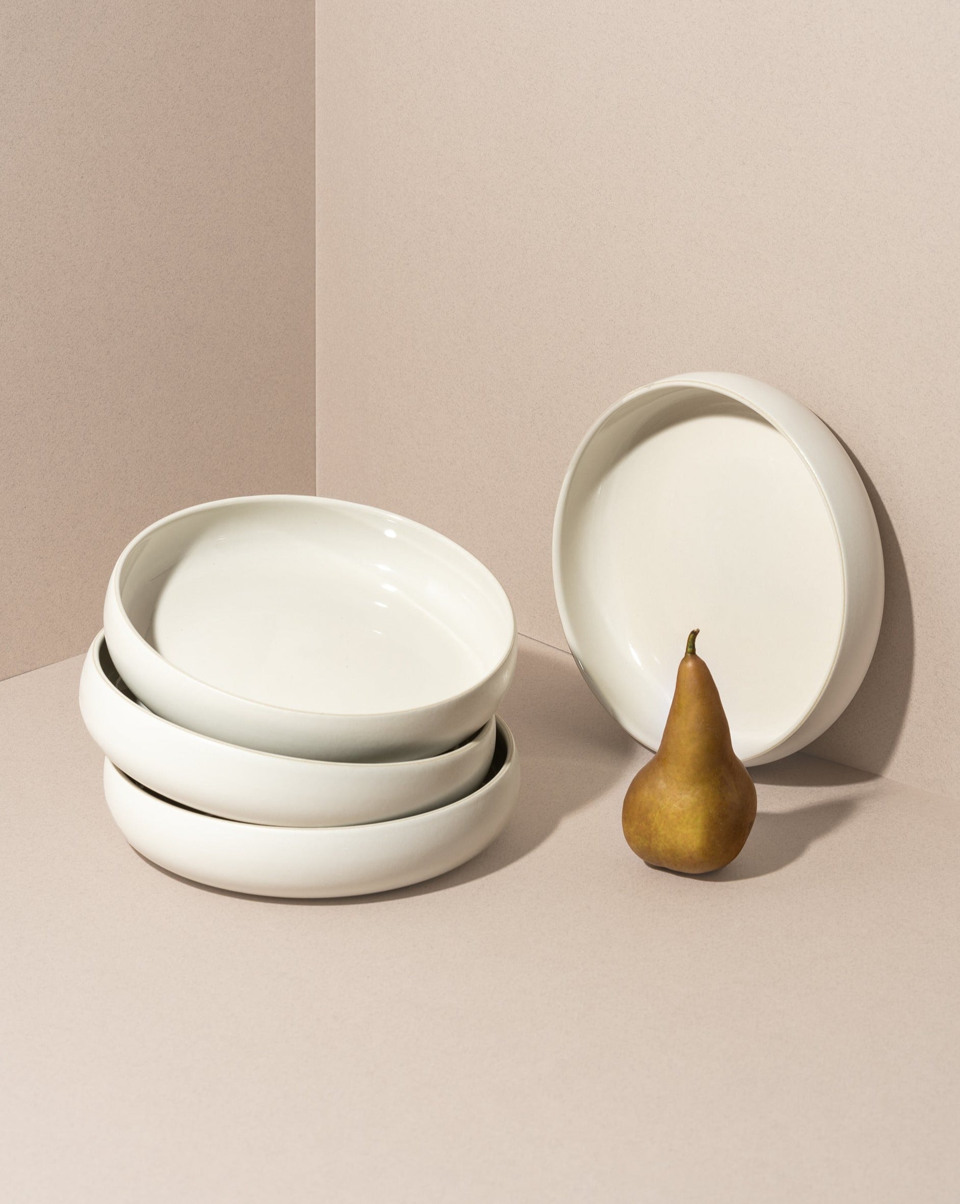 A beige stoneware pasta/salad plate with a contemporary round shape, displayed in a stacked arrangement with a white vegetable on the side.