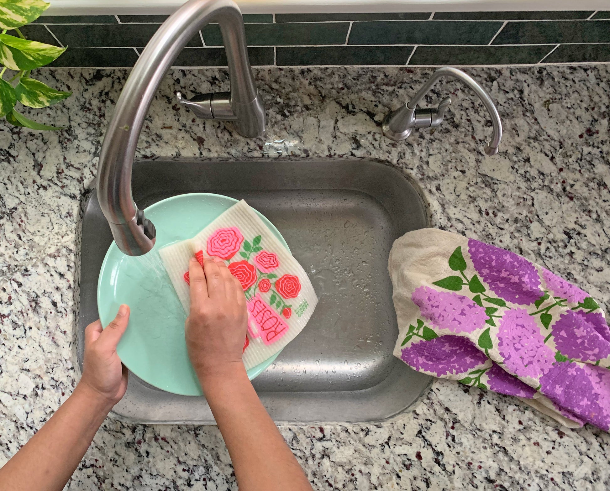 A woman washes a dish with a Roses  flower sponge cloth, while a Lilac floral dish towel rests on the counter.