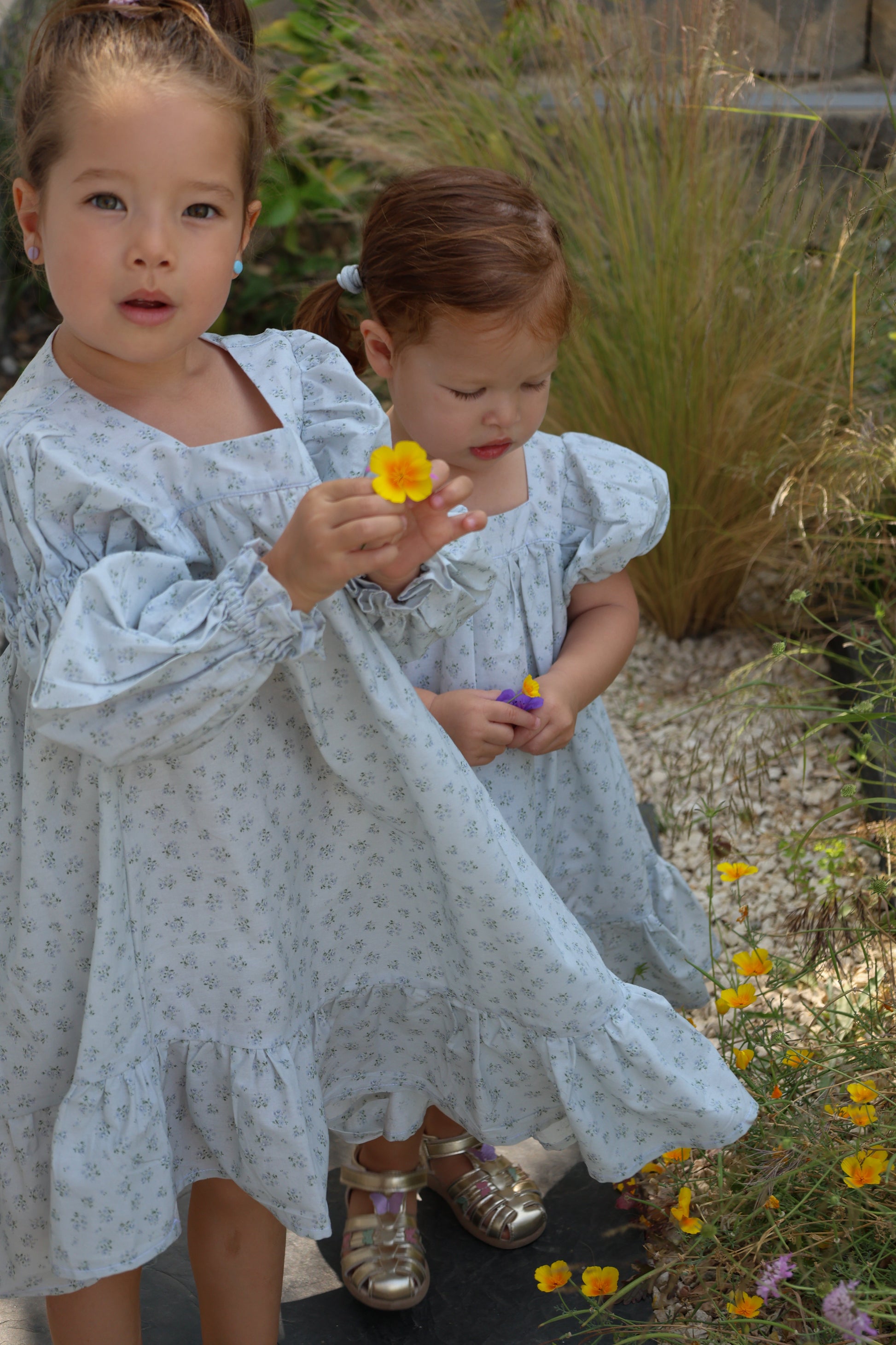 Two young girls in light blue dresses standing in a garden with flowers.