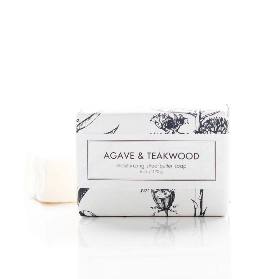 A neatly packaged bar of "Agave & Teakwood" moisturizing shea butter soap is placed on a white surface. The white wrapper features delicate botanical illustrations, and the soap itself is partially visible behind the packaging.