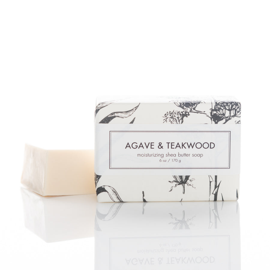 A bar of AGAVE & TEAKWOOD moisturizing shea butter soap wrapped in decorative paper with black illustrations. Another unwrapped soap bar is placed next to it. The product is placed on a white surface with a soft reflection below.