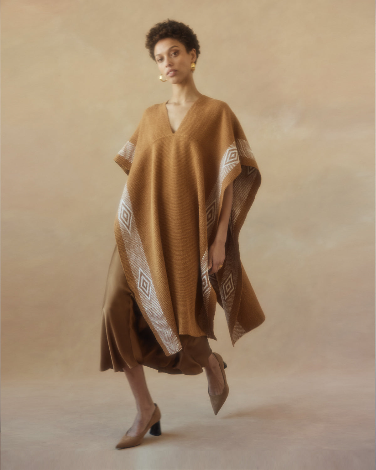 Model wearing a mid-length Manta Diamante poncho in gold with diamond patterns.