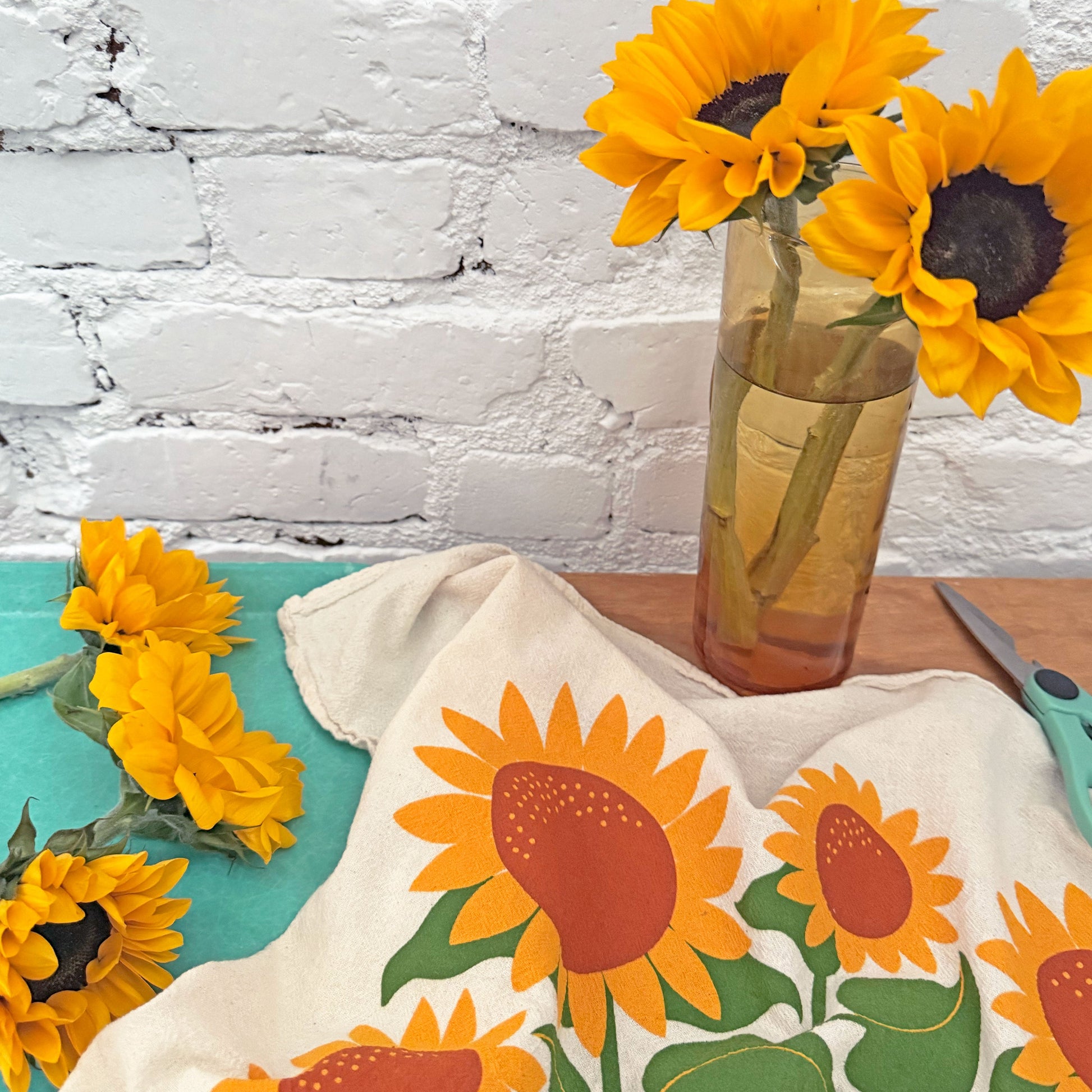 The Neighborgoods Sunflowers dish towel featuring a sunflower illustration, laid next to a vase of freshly cut sunflowers.