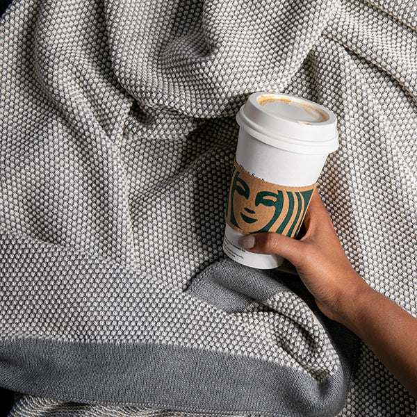 Person holding a Starbucks coffee cup while under a light grey and white textured cotton throw with a grey border.