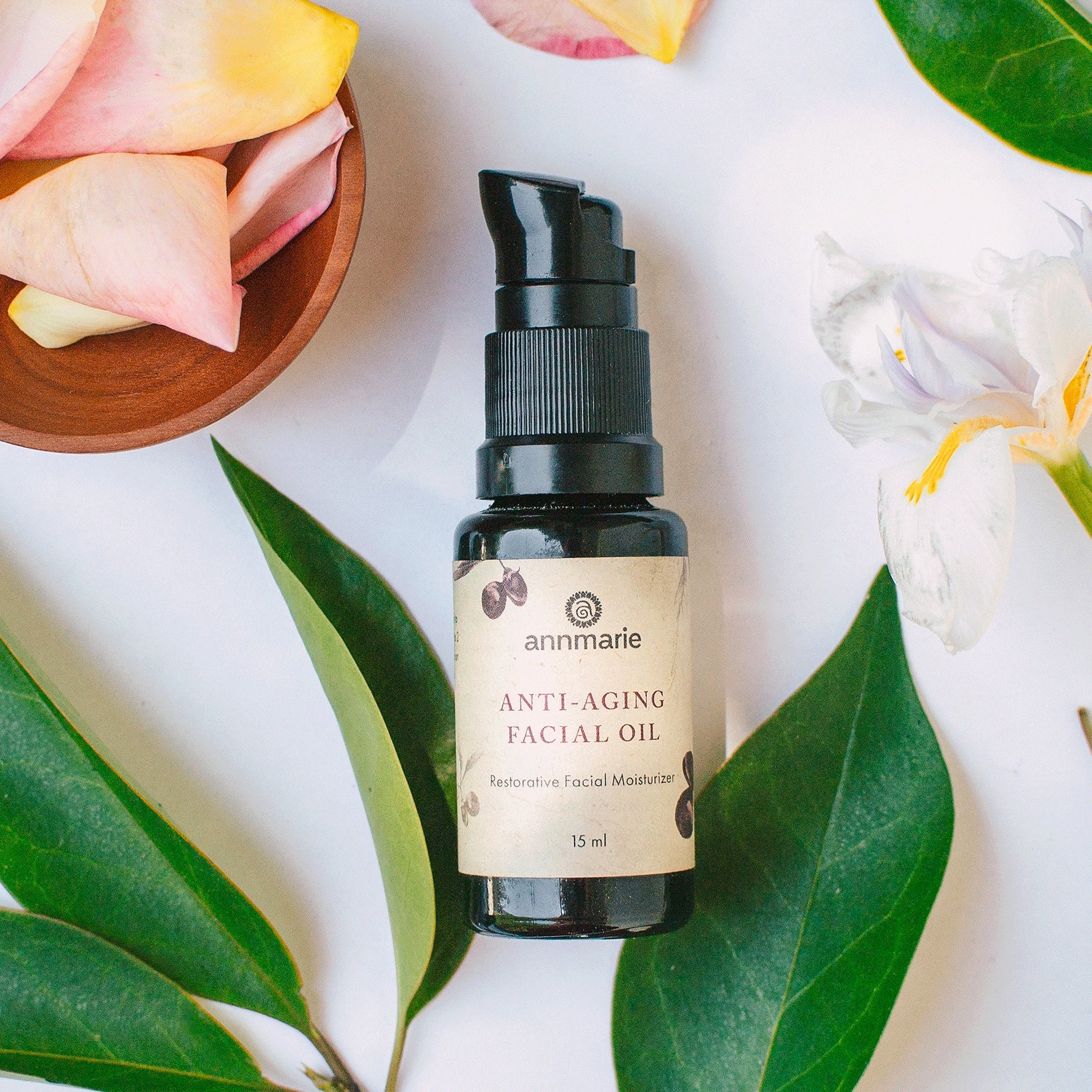 Bottle of Annmarie Skin Care Anti-Aging Facial Oil displayed with green leaves and soft flower petals representing natural skincare ingredients.
