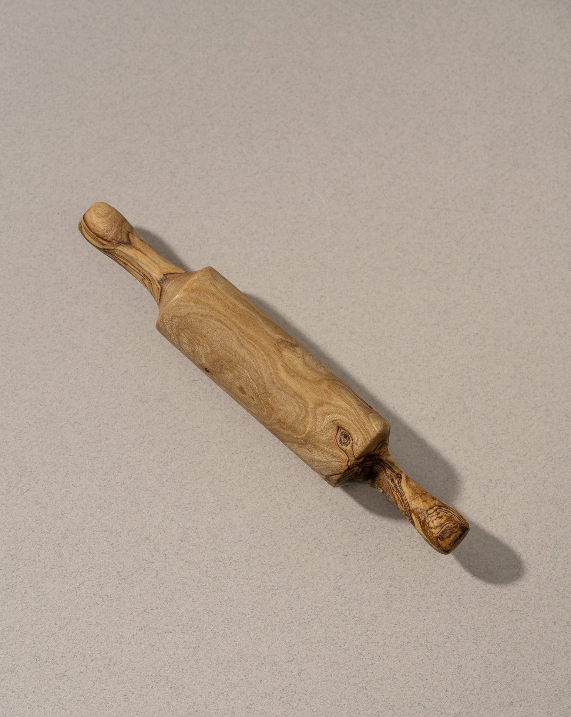 A single natural olive wood rolling pin displayed on an off-white background, highlighting its smooth finish, organic shape, and distinctive wood grain.