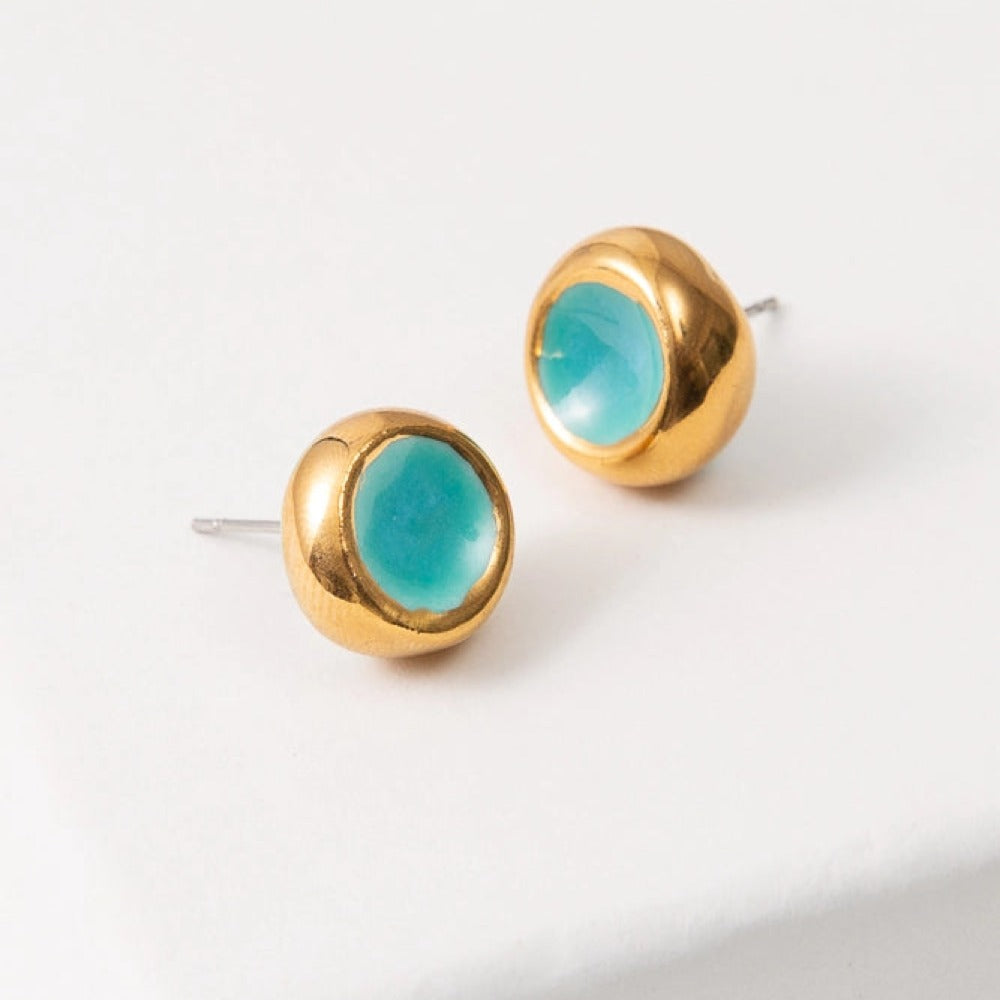 The Sculpted Porcelain Studs are composed of turquoise porcelain shaped into a small circle that is concave in the center. Shining gold luster coats the exterior rim of the studs, serving as a border.