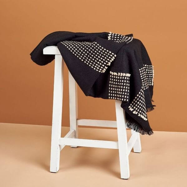 Walnut Black Yak With Hand Knotted Merino Stripes Handloom Throw draped on a white stool against a warm background.