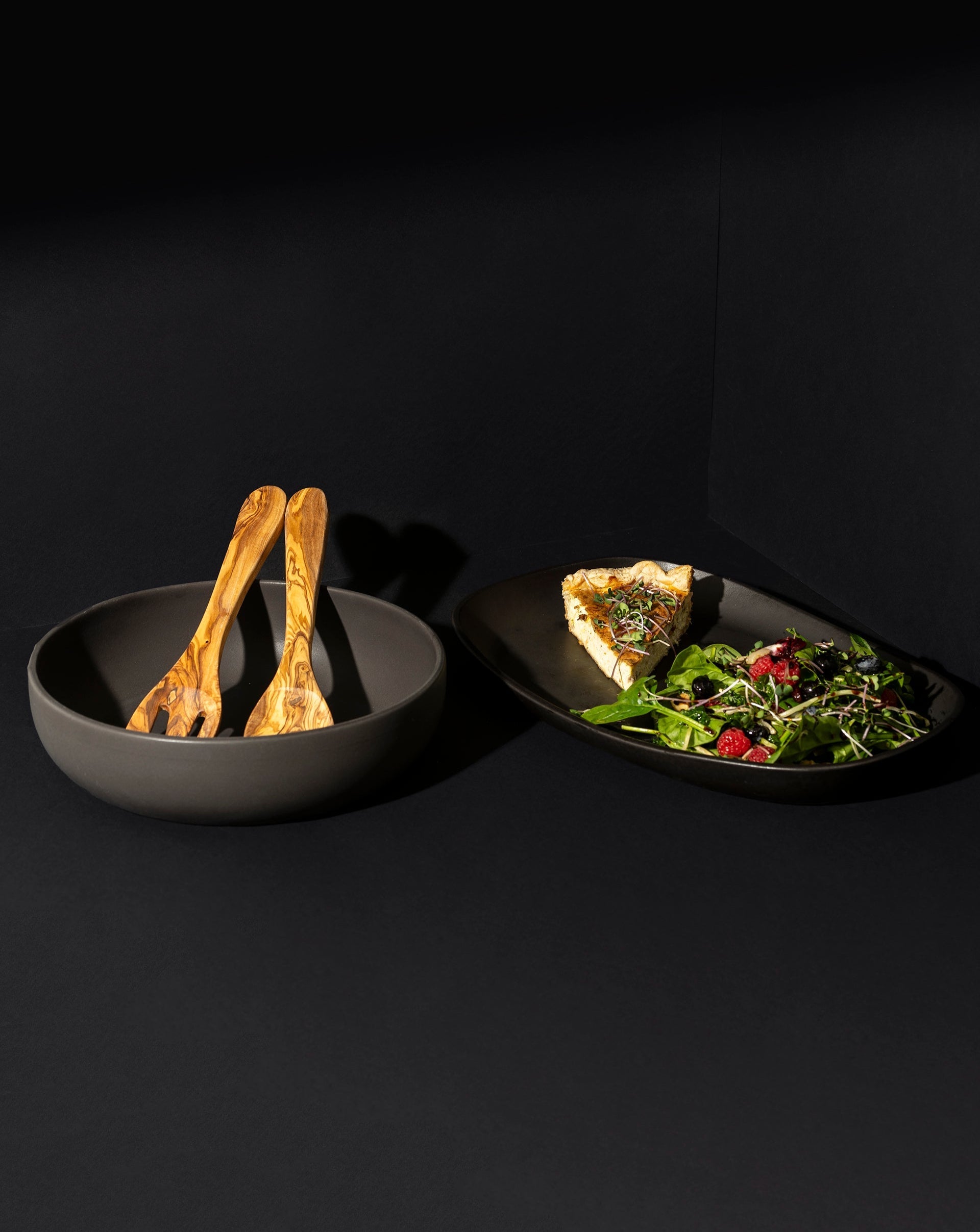 A white ceramic serving platter and bowl set with an olive wood serving set in the background, displayed with salad in the platter and garlic in the bowl.