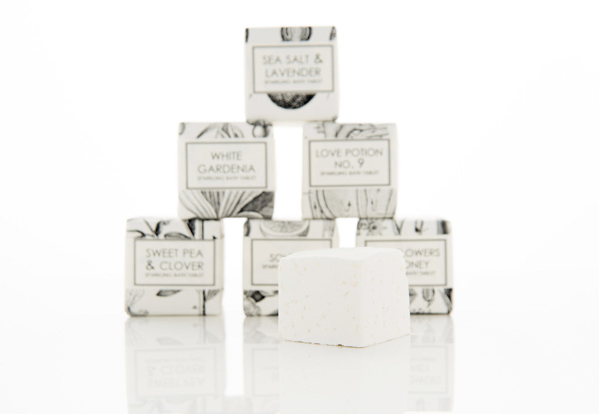 A pyramid of six boxed soaps is displayed against a white background. In front of the pyramid is an unwrapped white soap. The visible labels on the boxes read "SEA SALT & LAVENDER," "WHITE GARDENIA," "LOVE POTION NO. 9," "SWEET PEA AND CLOVER," and "SUNFLOWER'S HONEY.